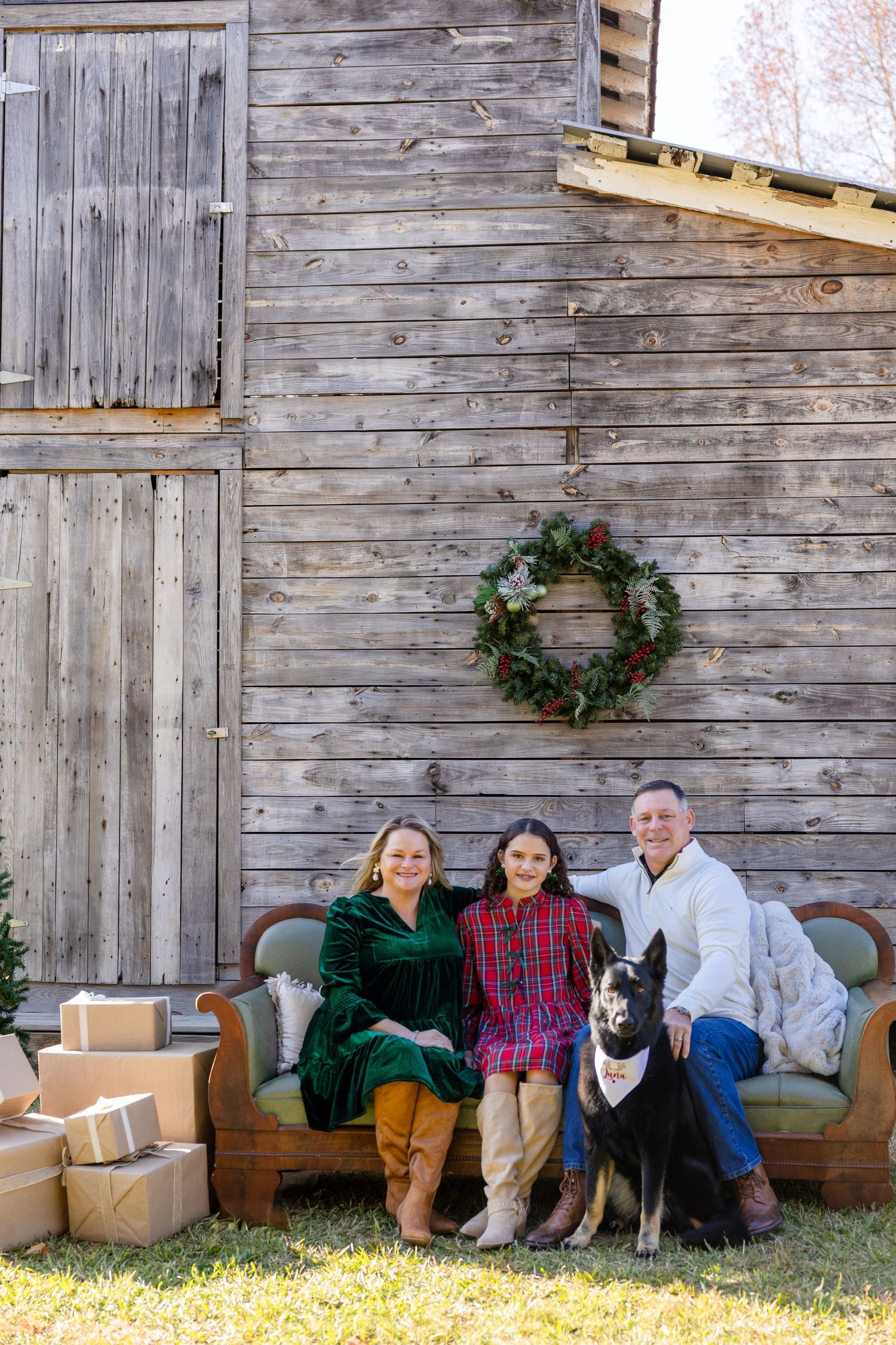 Family of three with black dog posing on outdoor sofa beside rustic barn with Christmas wreath and gifts.