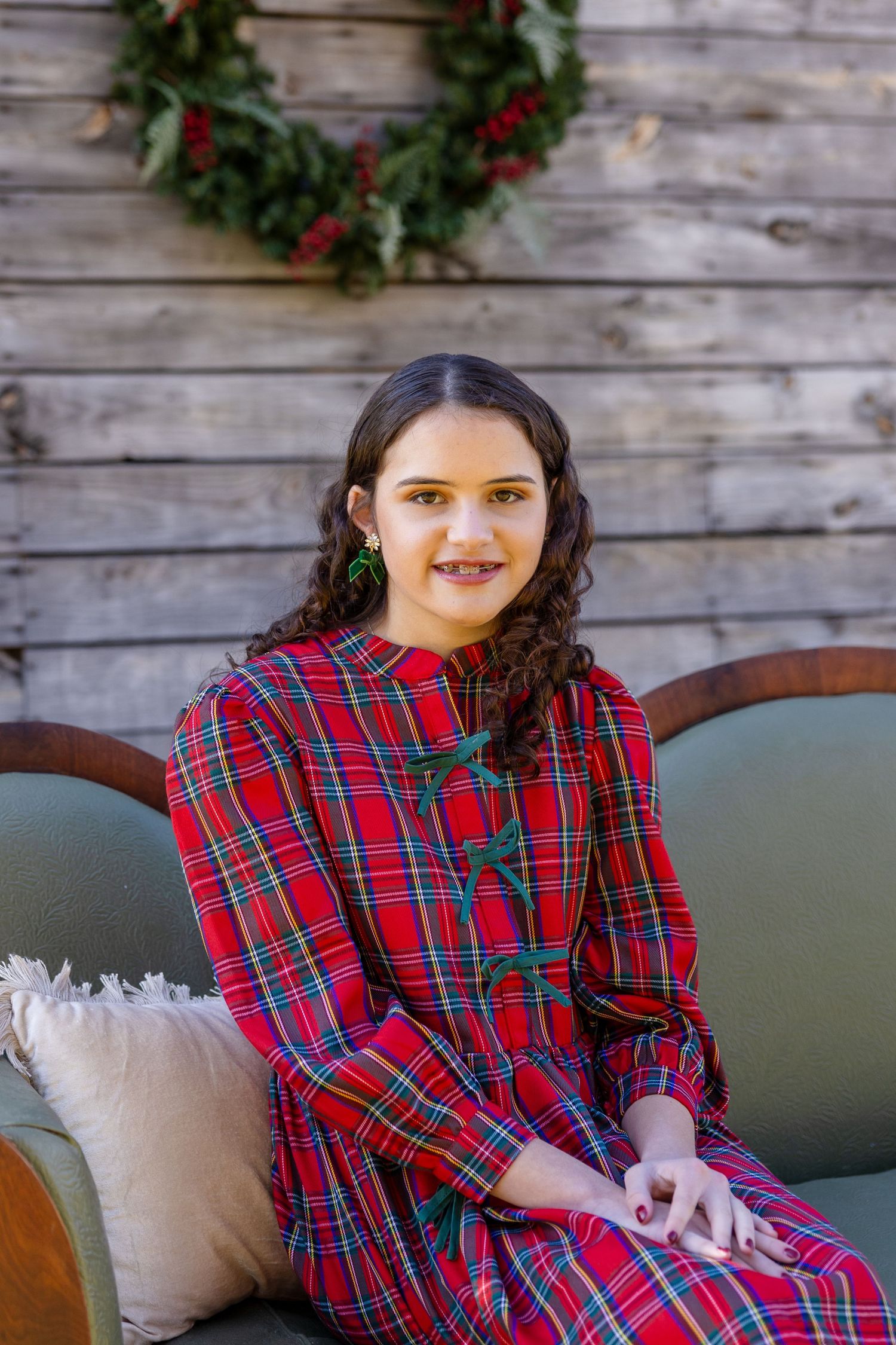 Young girl in red plaid dress sitting on sofa with Christmas wreath backdrop, smiling for holiday portrait.