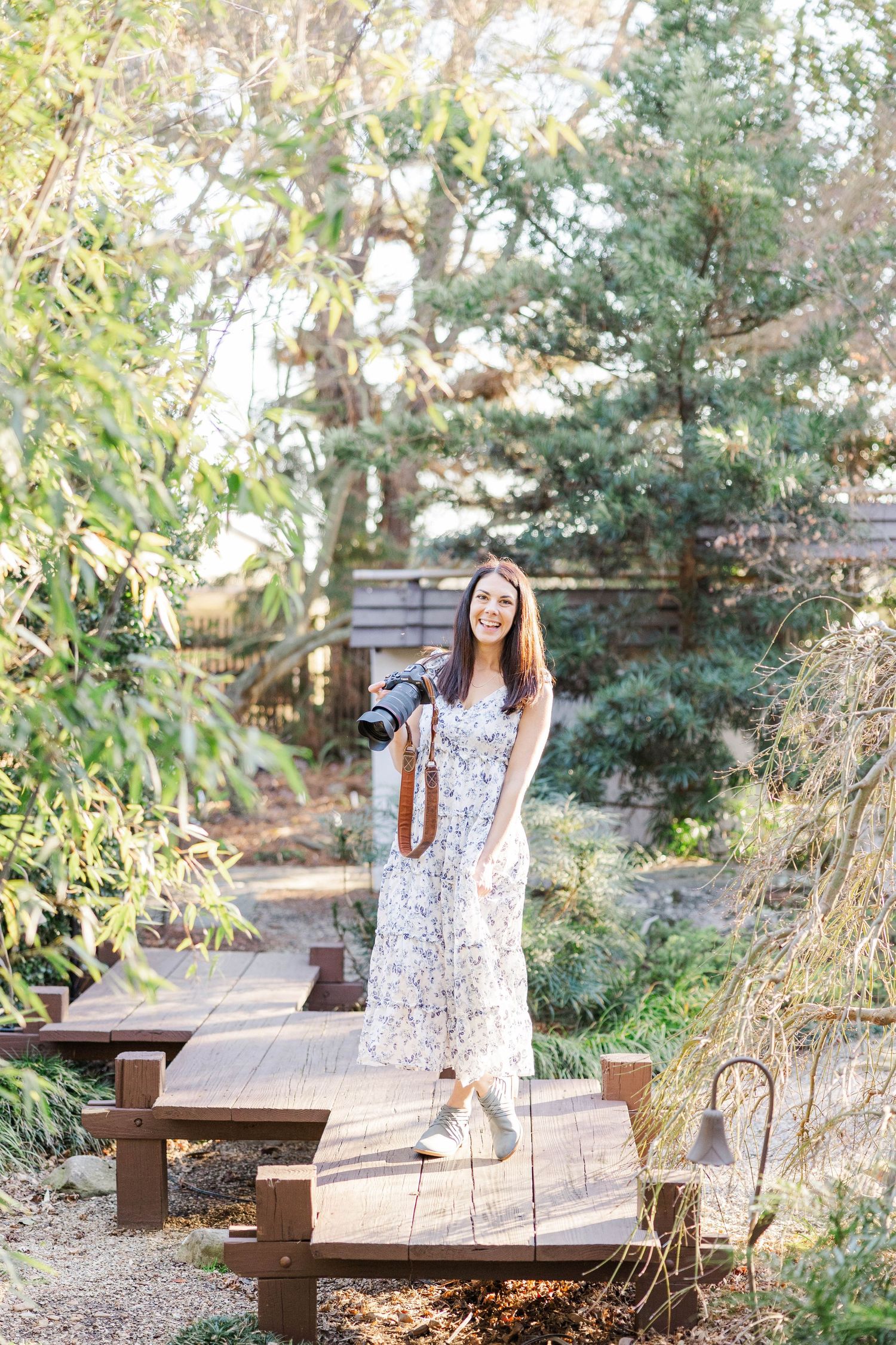 A Raleigh family photographer stands with her camera in a garden, wearing a white and blue dress.