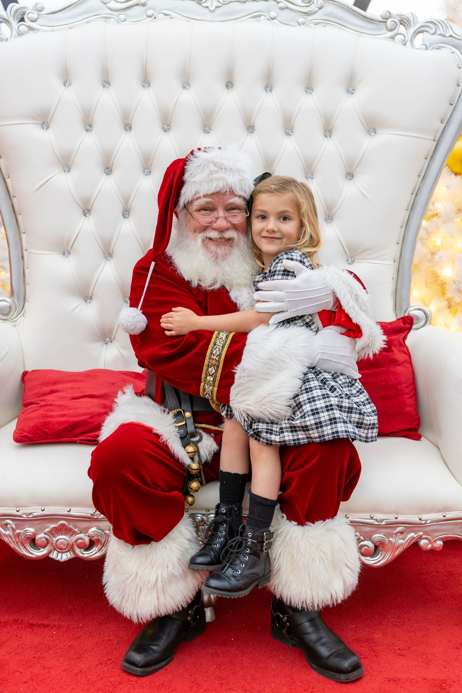 A young girl hugs Santa Claus sitting on a white throne chair with red cushions during a Christmas photo session.