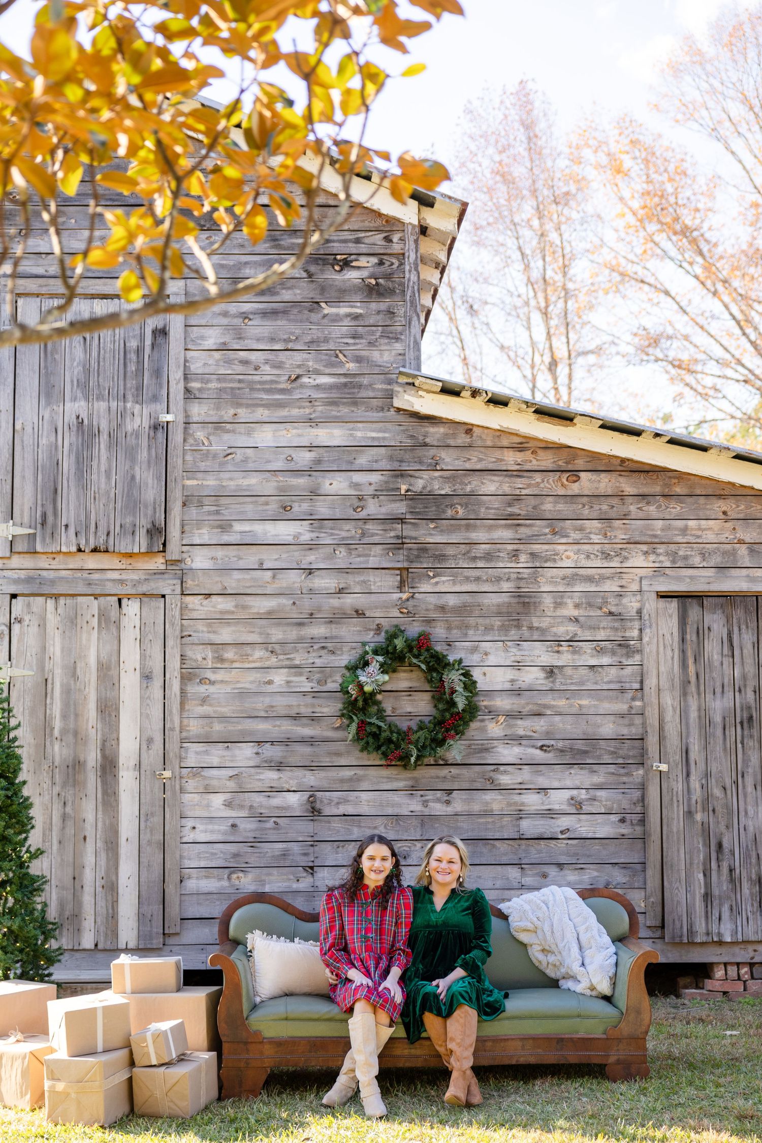 Two women sitting on a vintage sofa outdoors by a rustic barn with a Christmas wreath and gift boxes.