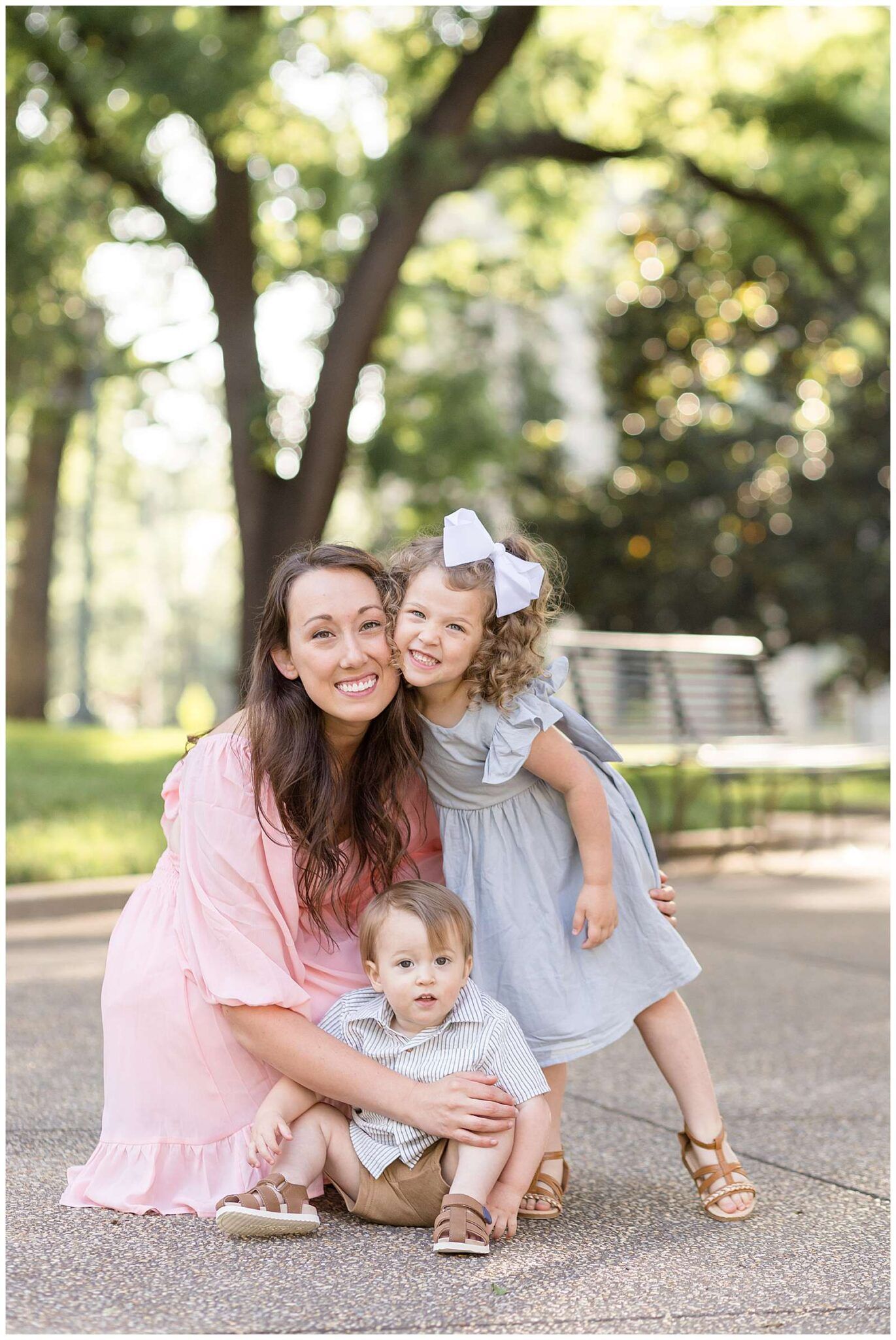 A smiling mother in a pink dress poses outdoors with her toddler son and young daughter in a dress and white bow.