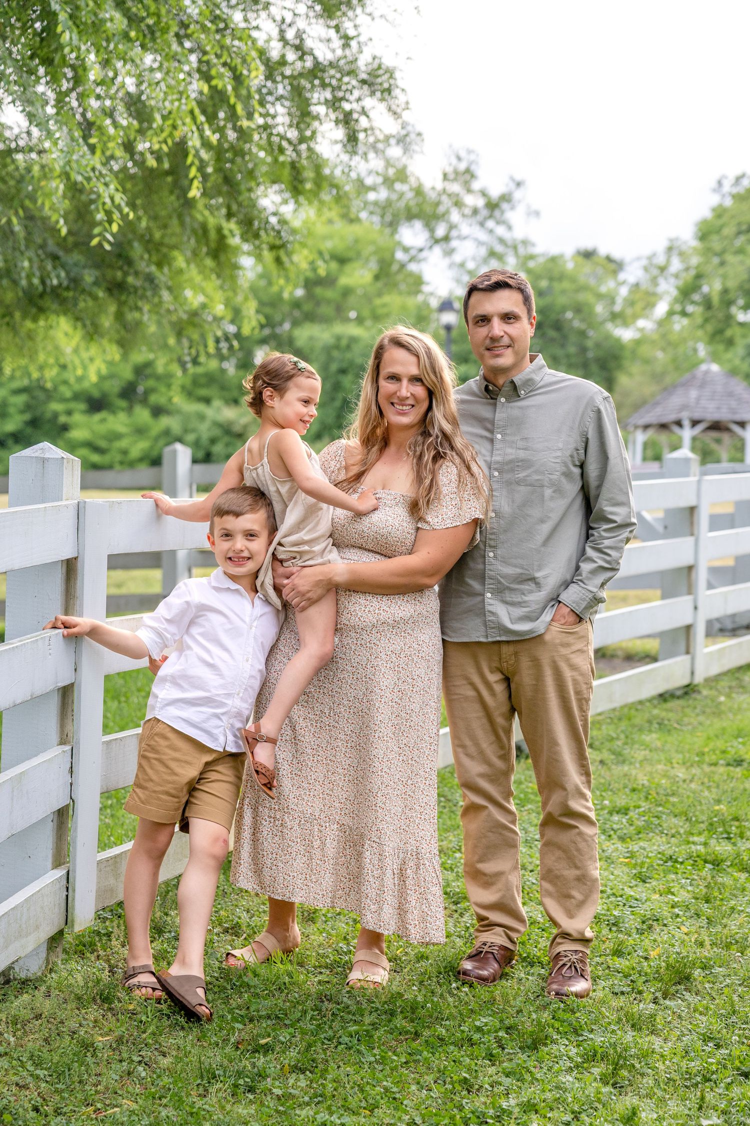 A family shares playful moments together along a white fence on a grassy hillside in summer.
