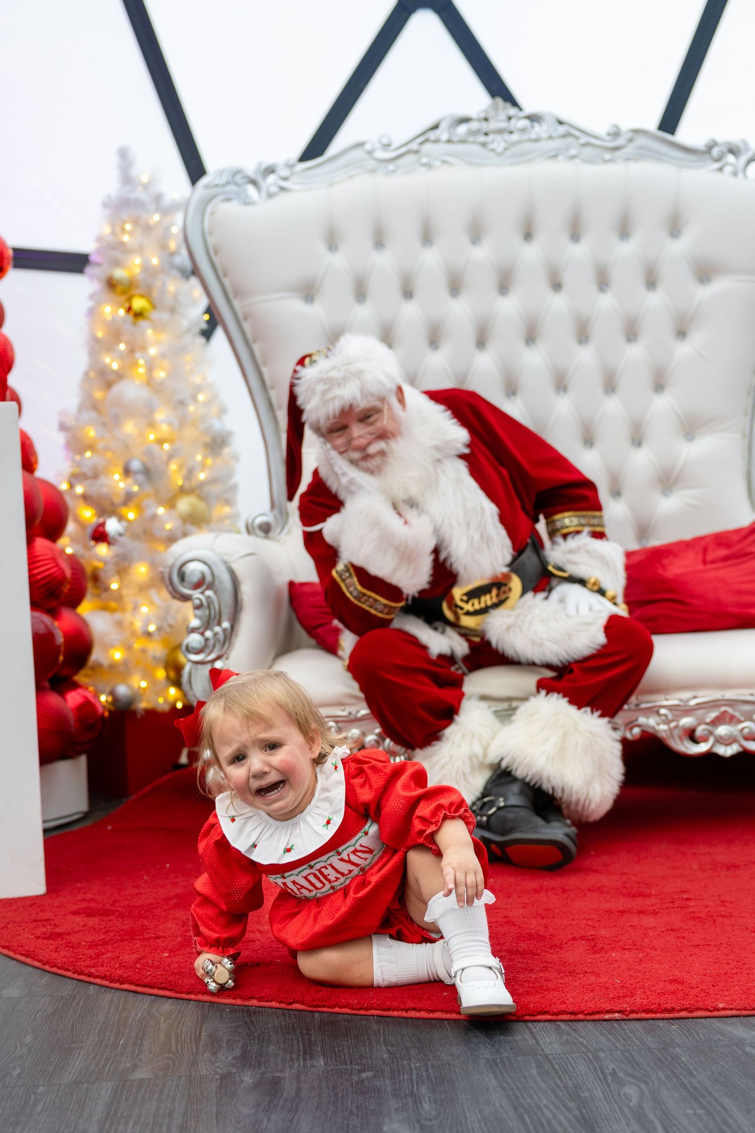 Crying toddler in red dress sits on floor before Santa Claus on white throne beside decorated Christmas tree.