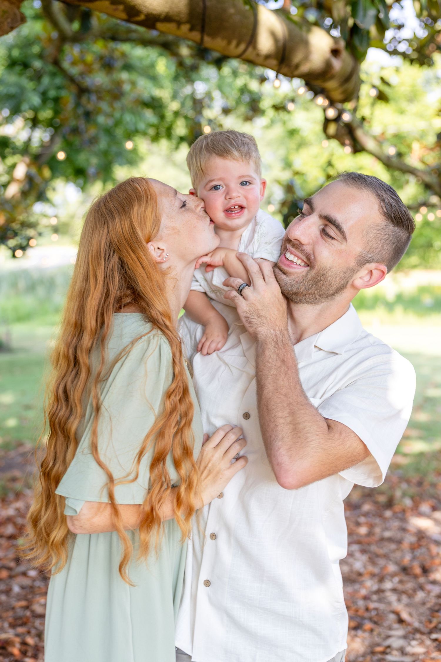 Happy family with redhead mom kissing toddler boy held by smiling dad outdoors under green tree - Raleigh NC family photography