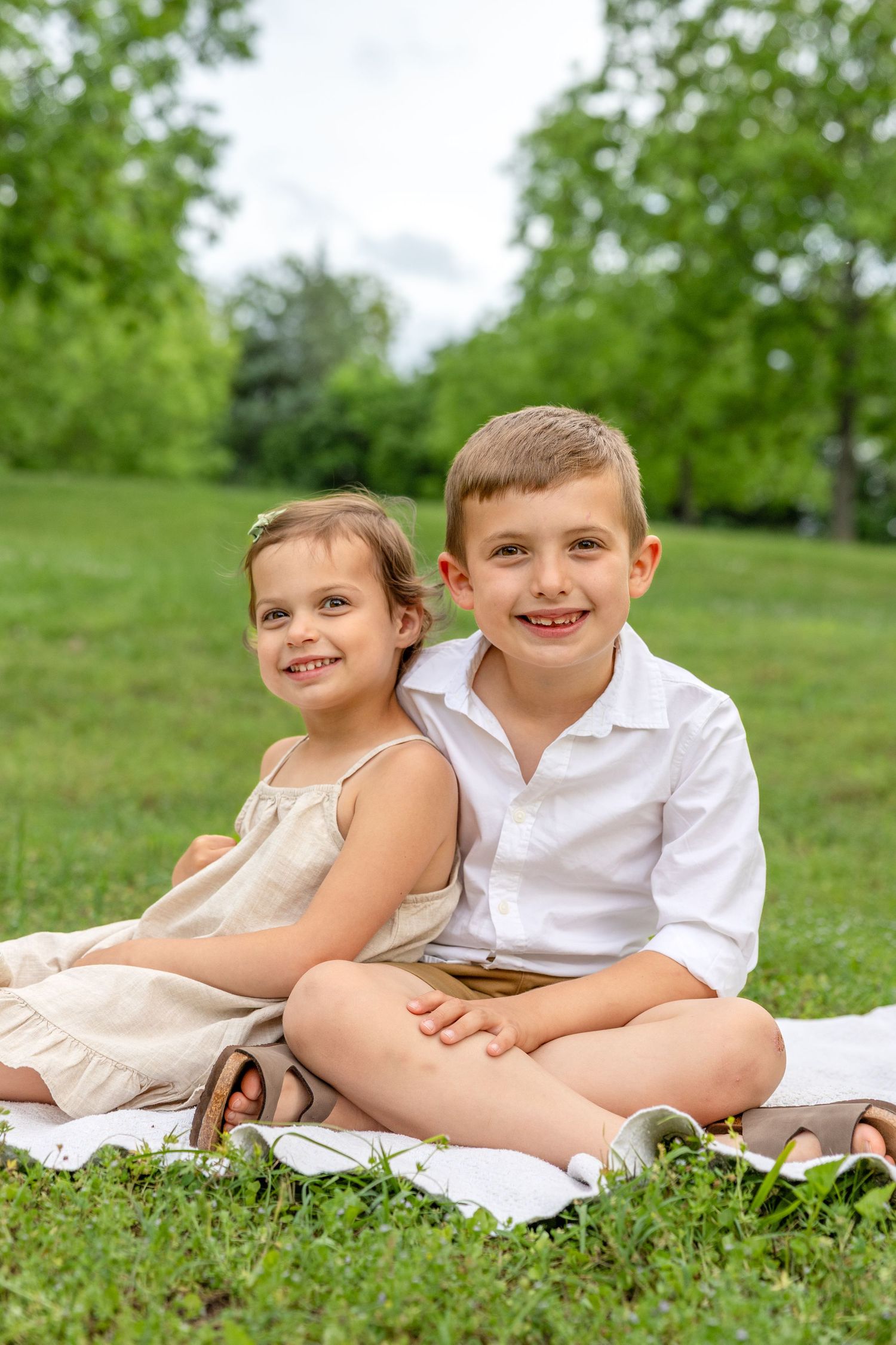 Two children in light colored clothing sit together on grass during a spring outdoor photo session.