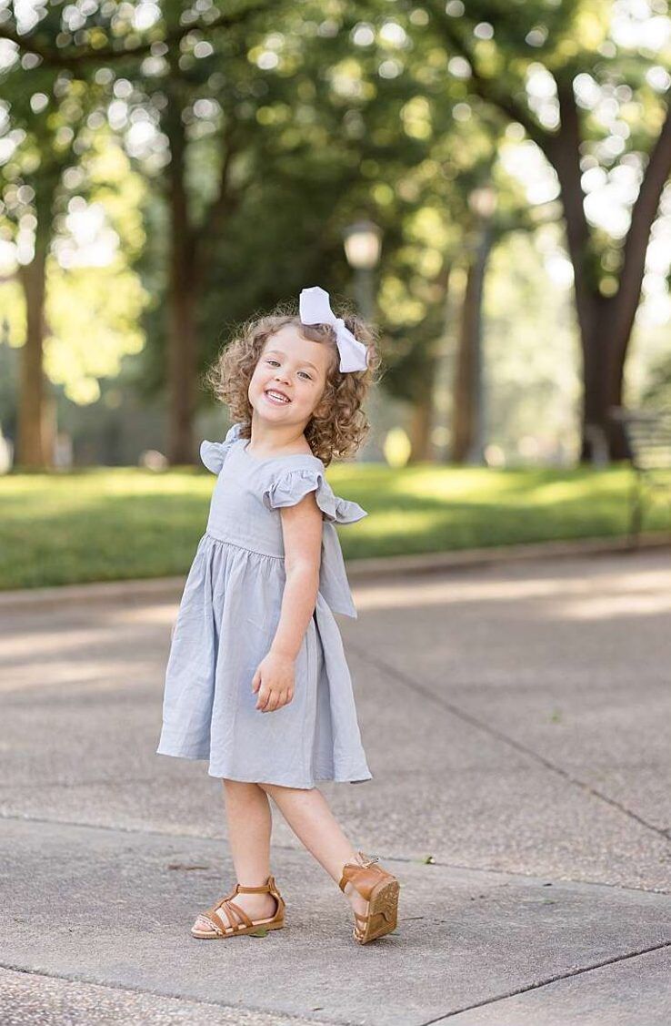 Smiling toddler girl in light blue dress with bow hair clip poses on park path in golden sunlight in Downtown Raleigh