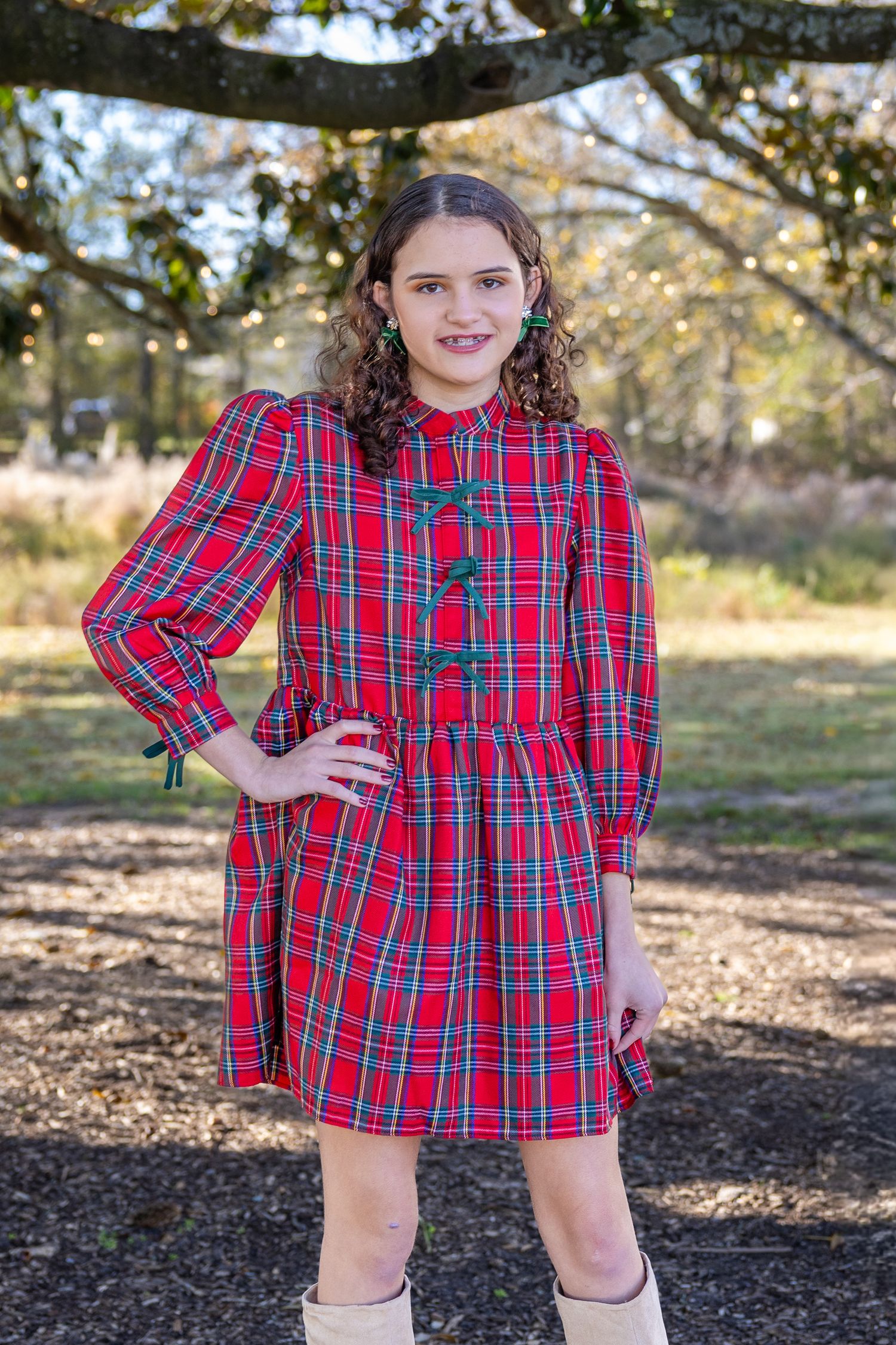 Young girl wearing a red tartan plaid dress with green buttons, posing outdoors under a tree.