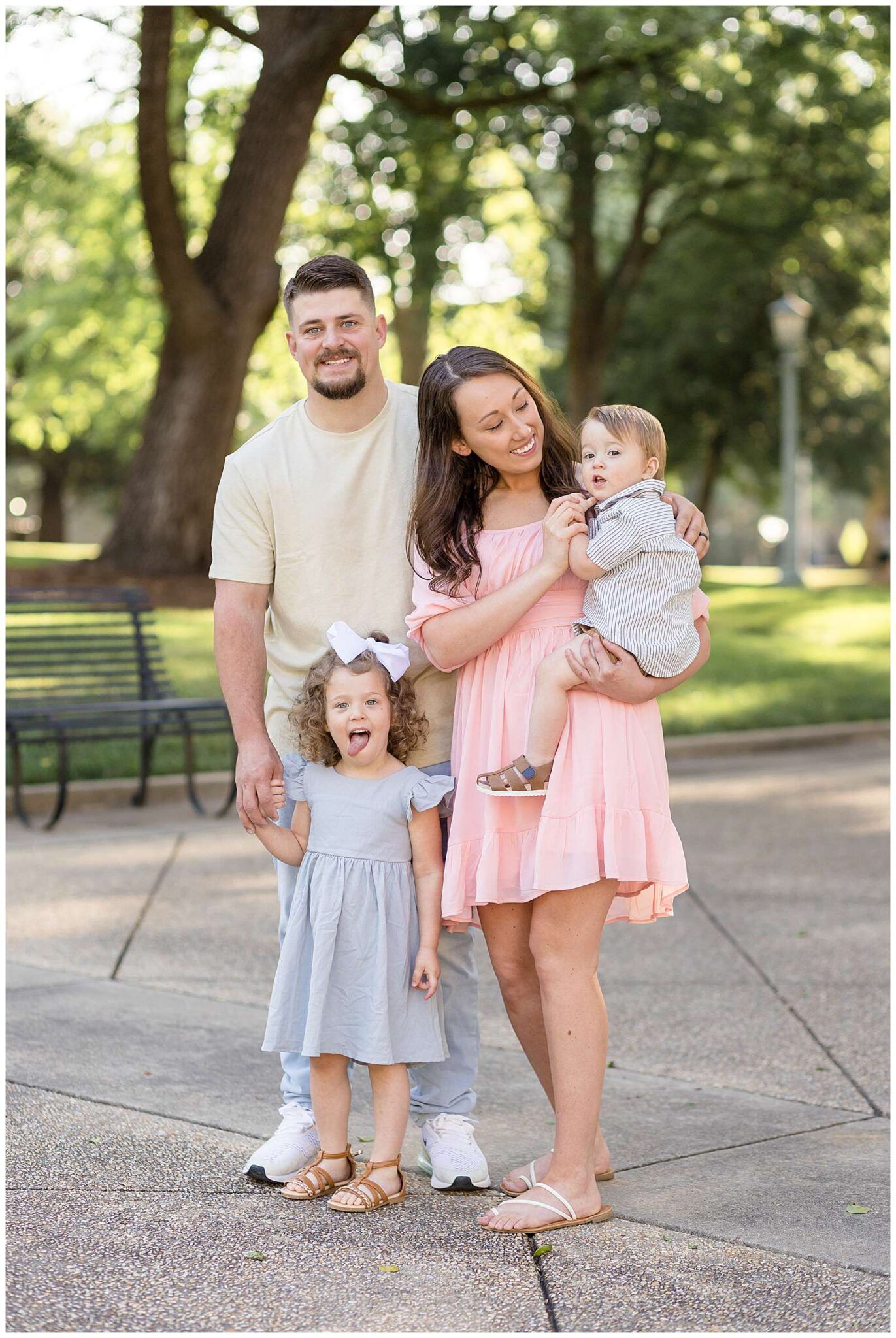 Happy family of four posing outdoors in a park, parents with toddler girl and baby boy in pastel outfits.