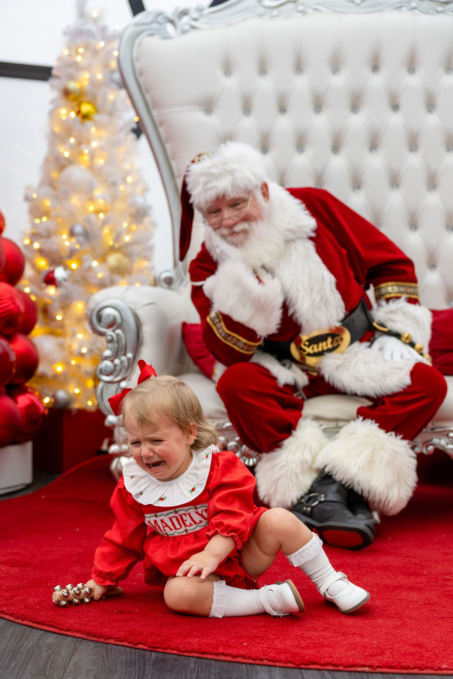 Crying toddler in red dress sits on floor during Santa Claus photo session in Cary, NC at Fenton.