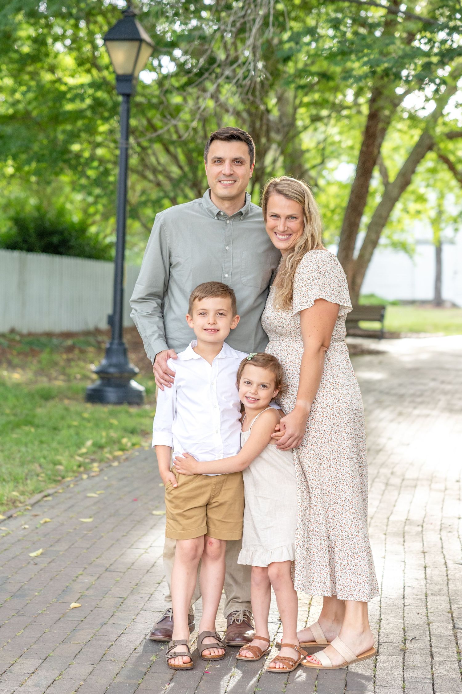 A family poses together on a brick pathway surrounded by green trees and a vintage lamppost on a sunny day.