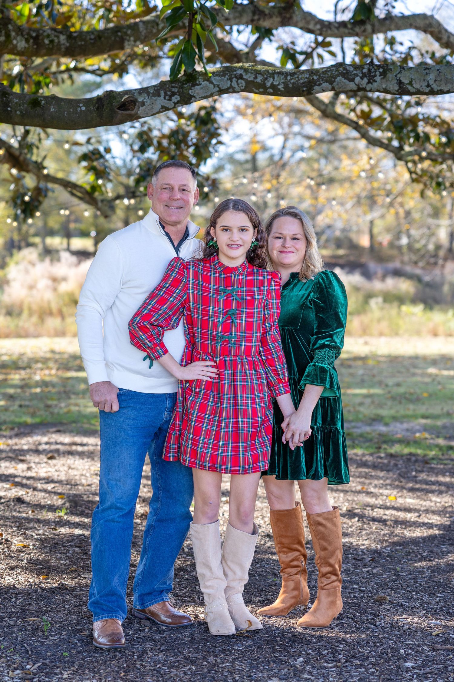 Family of three posing outdoors in holiday attire under oak tree, girl in plaid dress stands by parents in festive green and white.