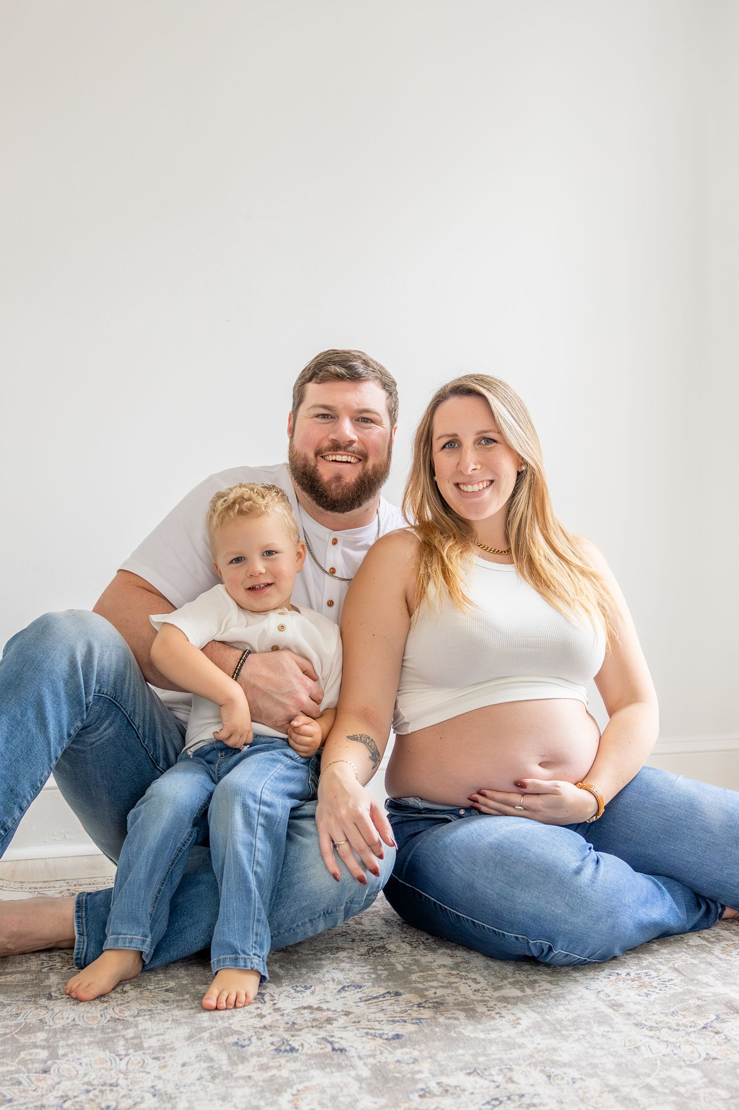 Smiling pregnant woman sits with bearded husband and toddler child in matching white tops and jeans for a family maternity photo.