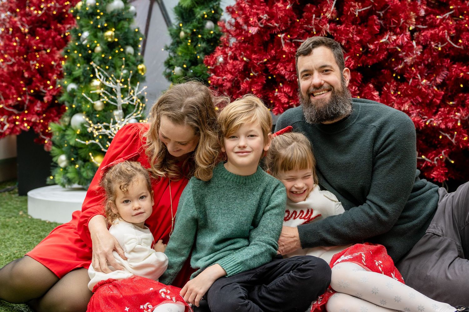 Family of five posing for a Christmas photo in front of red holiday trees, wearing festive outfits.