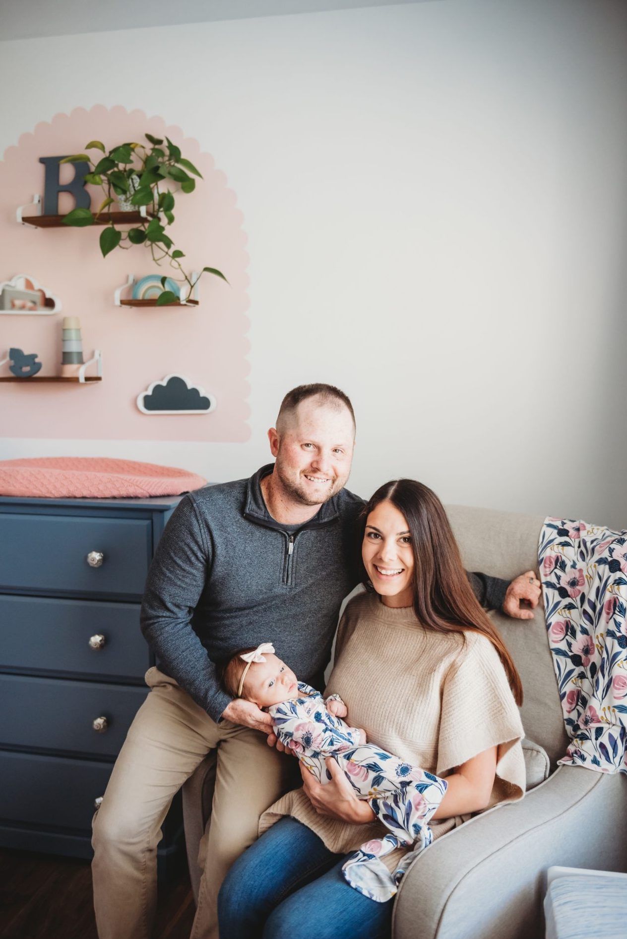 Family sits together in a nursery decorated with pink walls, floating shelves, and plants, alongside a dark dresser.