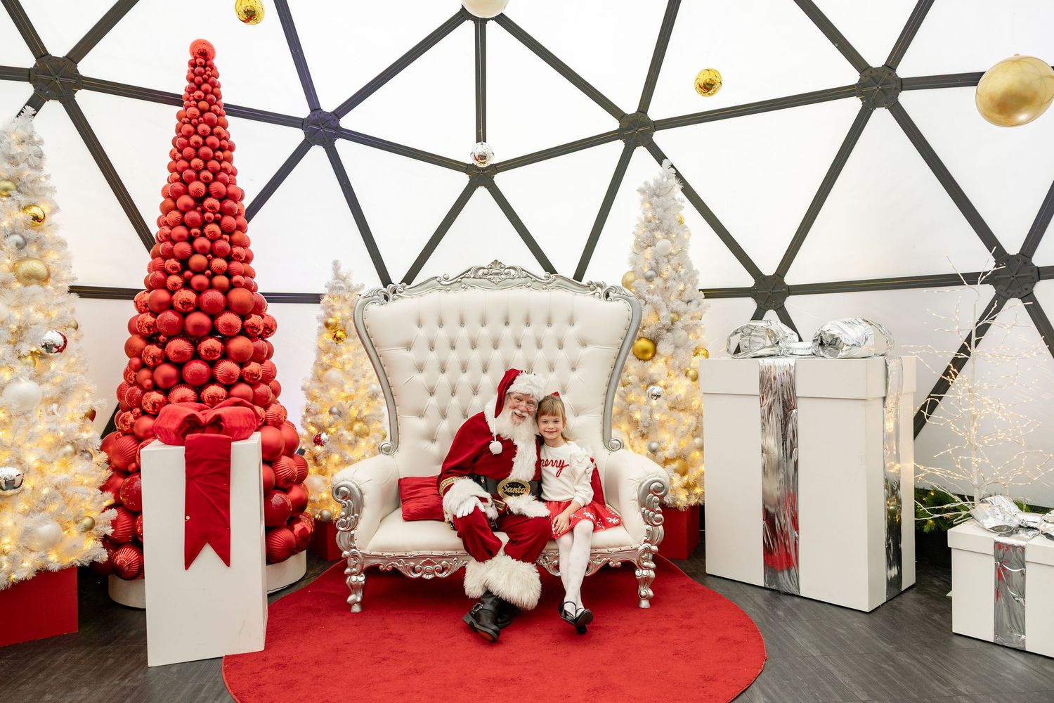 Child sits with Santa Claus on ornate white throne surrounded by red and white Christmas trees and gifts in festive dome.