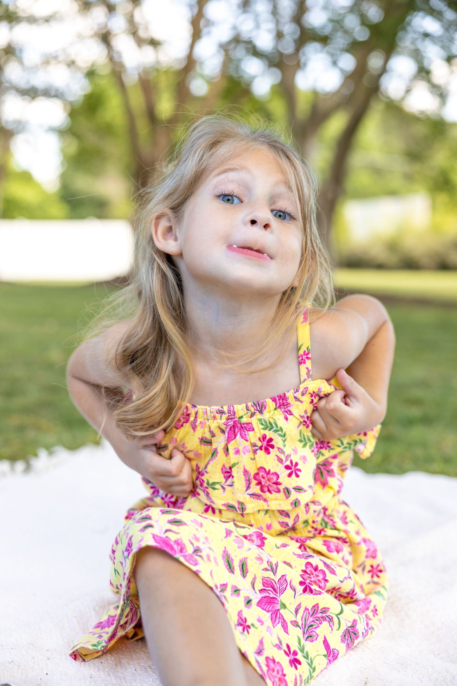 Young child in a yellow floral sundress poses playfully outdoors on a sunny summer day with trees in background.
