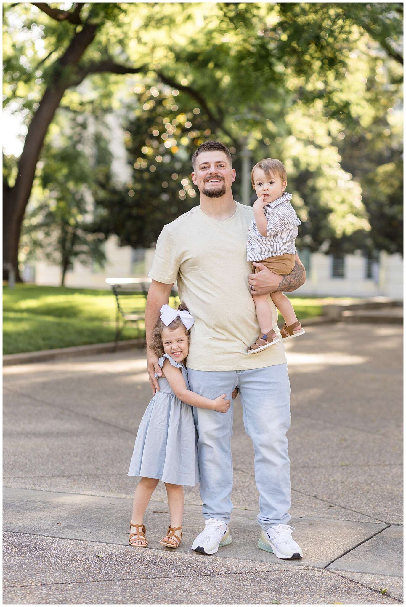A smiling Dad holds a baby while a young girl in a gray dress hugs his leg outdoors in a sunny park in Raleigh
