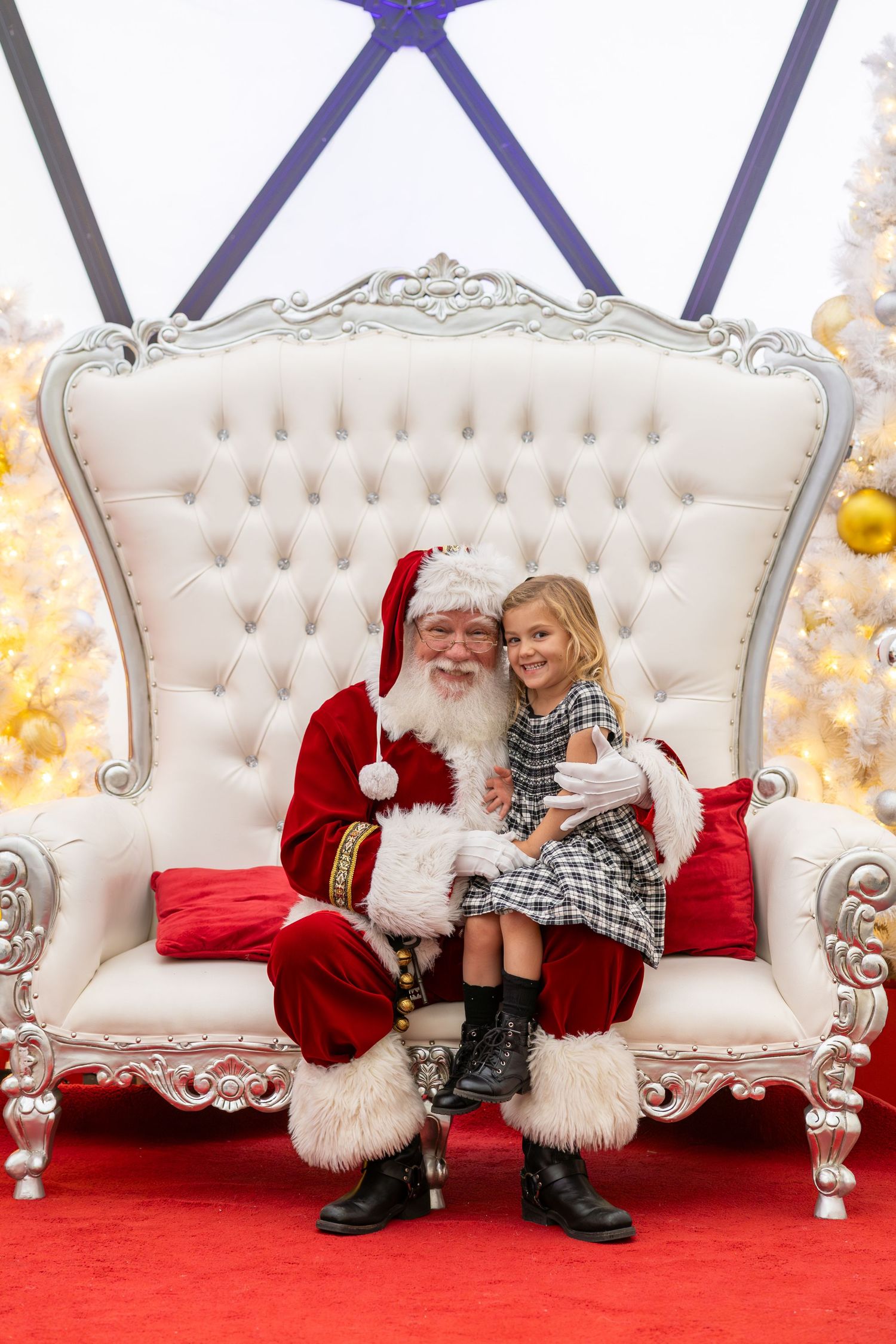 Young girl sits on Santa Claus's lap in a white throne chair surrounded by Christmas decorations.