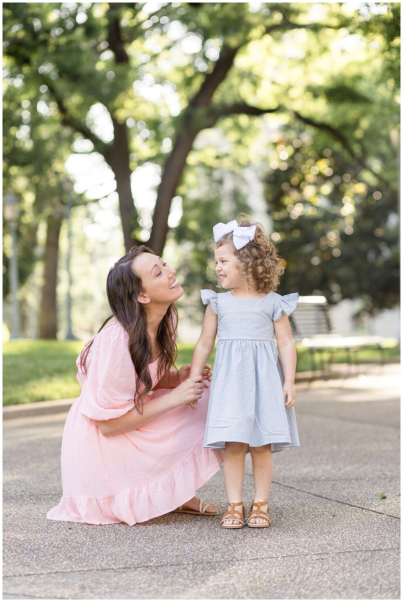A mother in a pink dress crouches beside her toddler daughter in a blue dress with a bow in a sunny park.