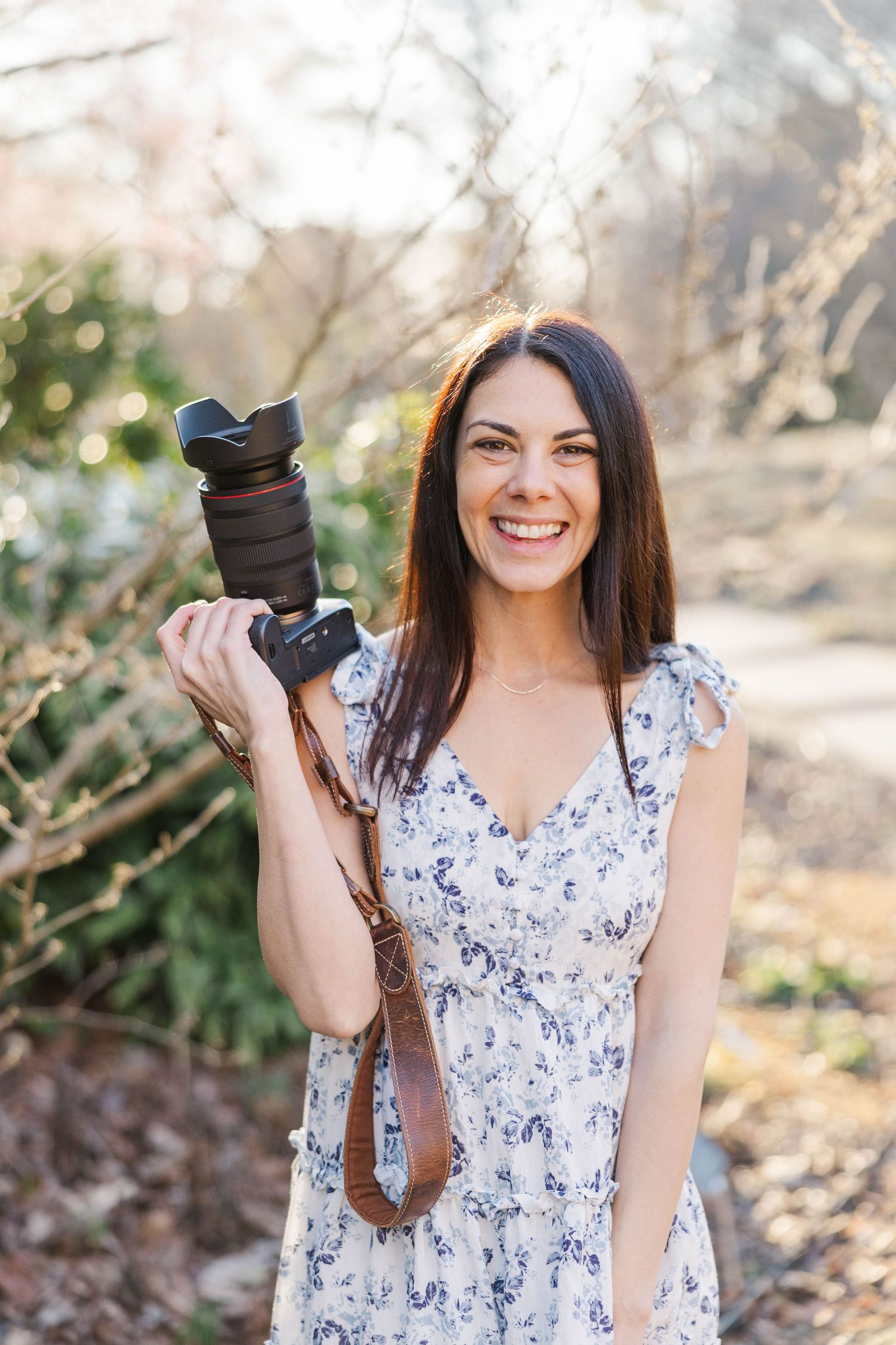 A Raleigh photographer in a floral dress holds a professional camera while standing outdoors in warm sunlight.