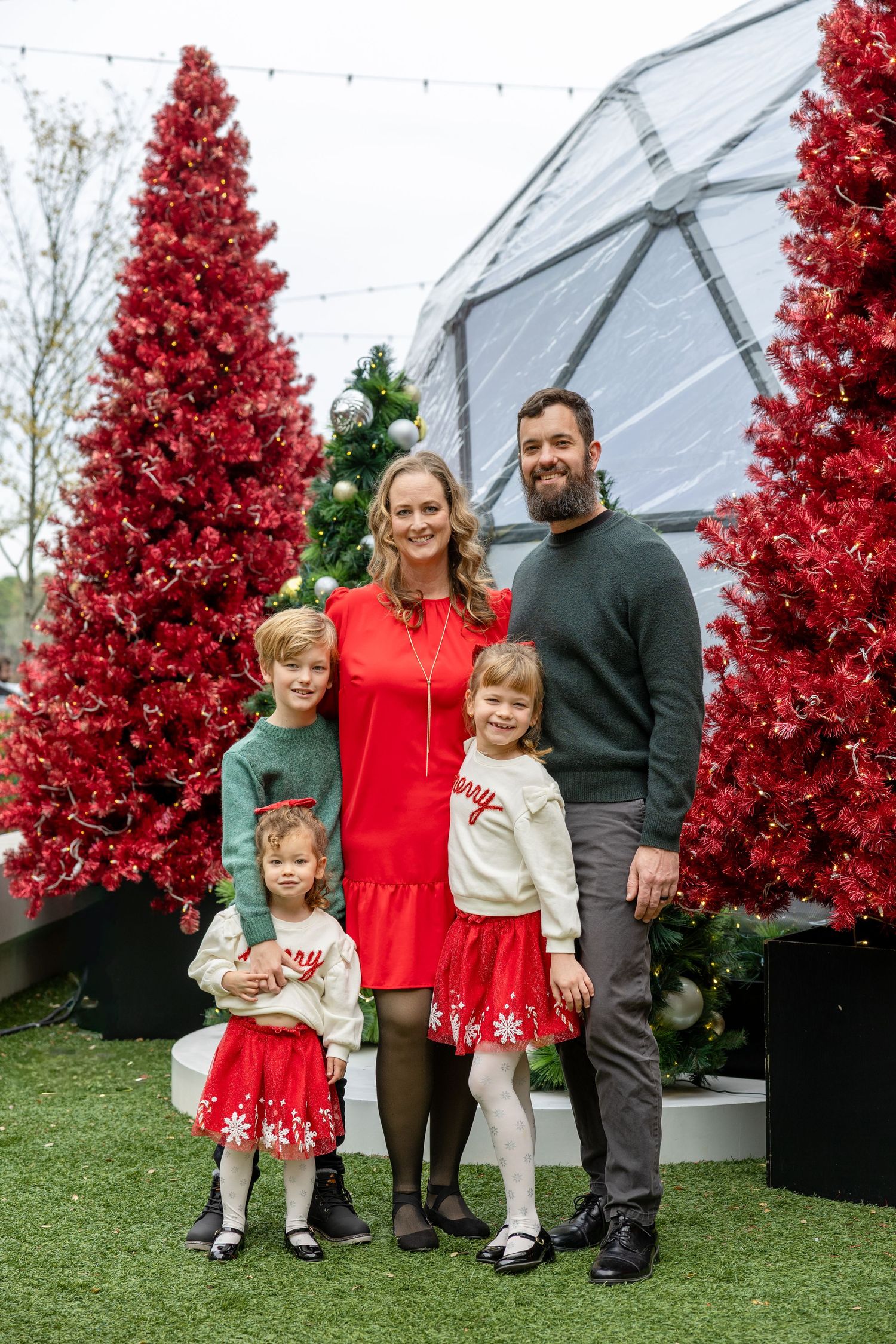 Family of five posing in front of red Christmas trees in festive holiday outfits outdoors.