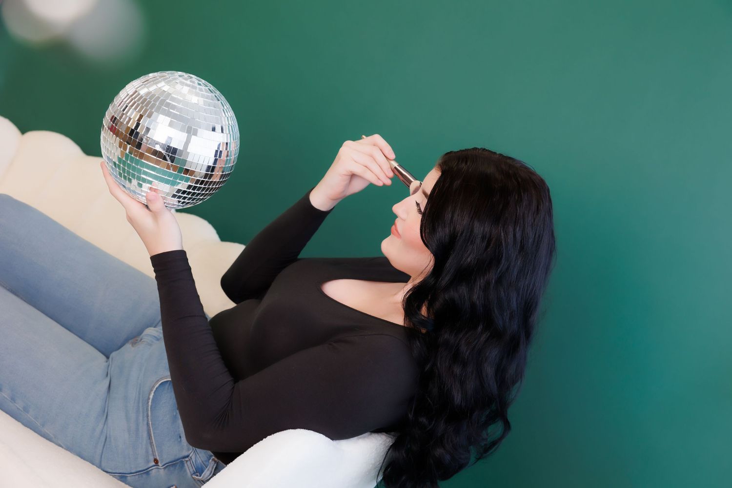 Person in black top lying on white surface next to disco ball against turquoise background.