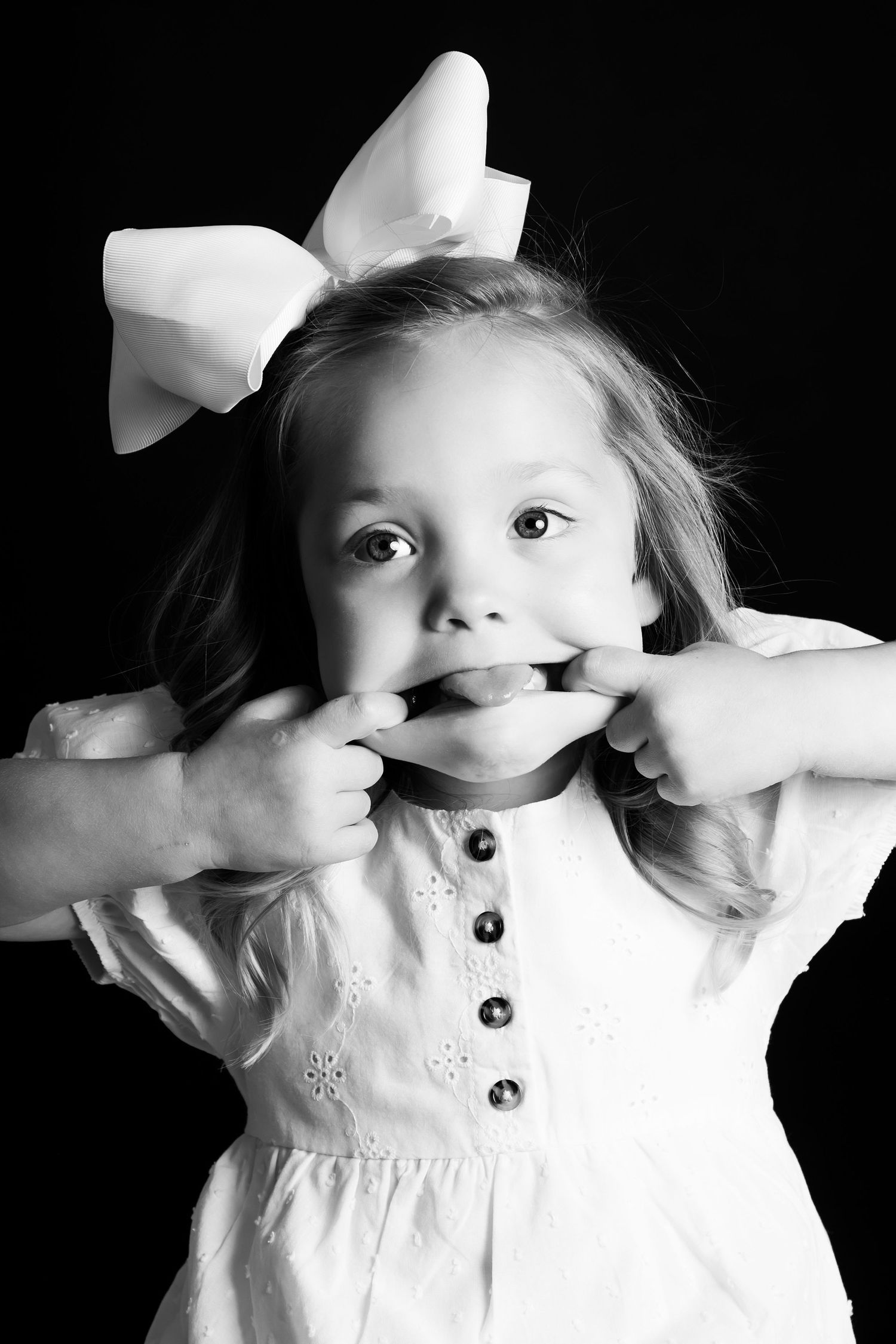 Black and white portrait with white bow showing silly face and pulling cheeks.