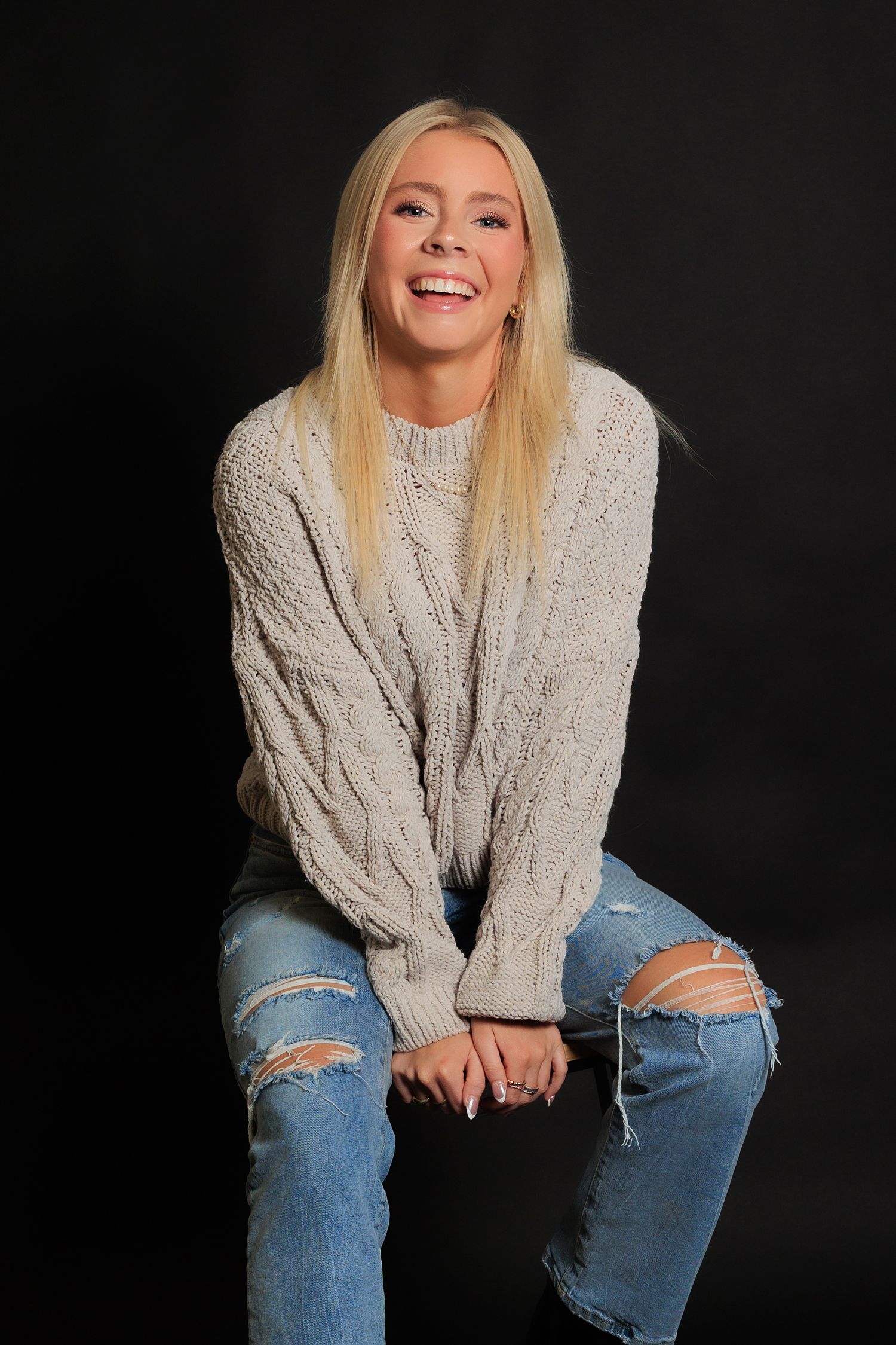 Portrait in beige cable knit sweater and ripped jeans against dark background with joyful expression.