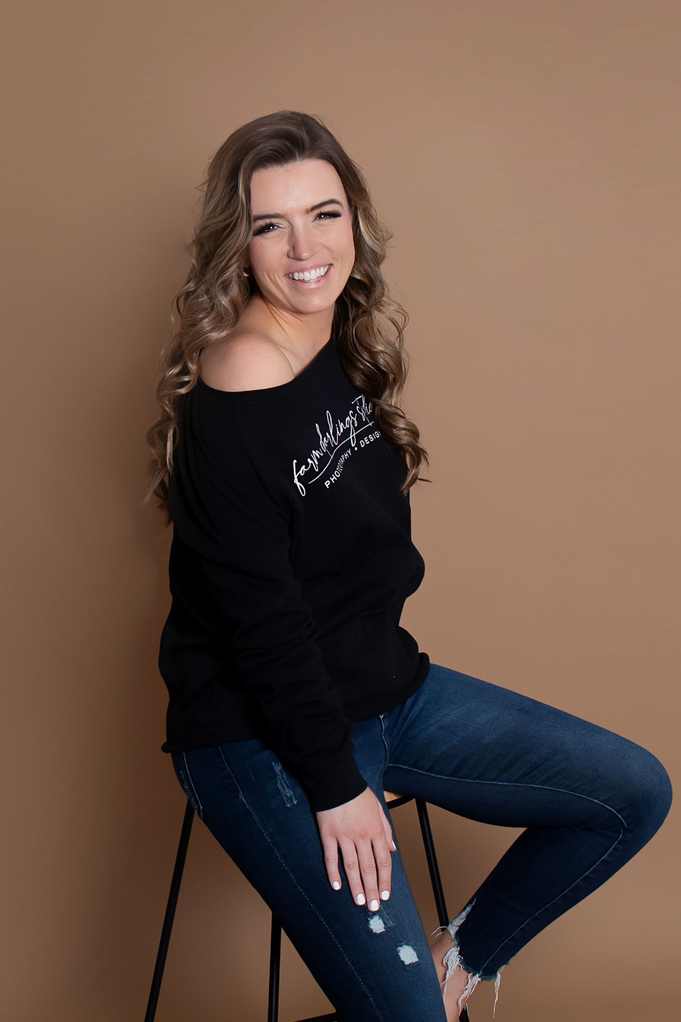Studio portrait in black sweater and jeans against brown background with warm smile.