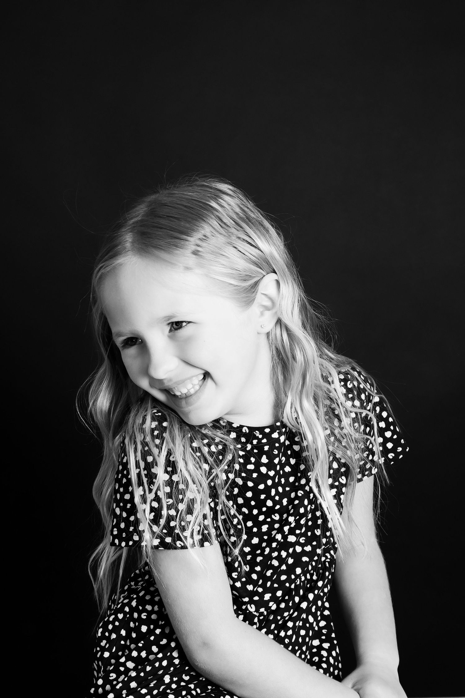 Black and white portrait showing shy smile in polka dot dress against dark background.