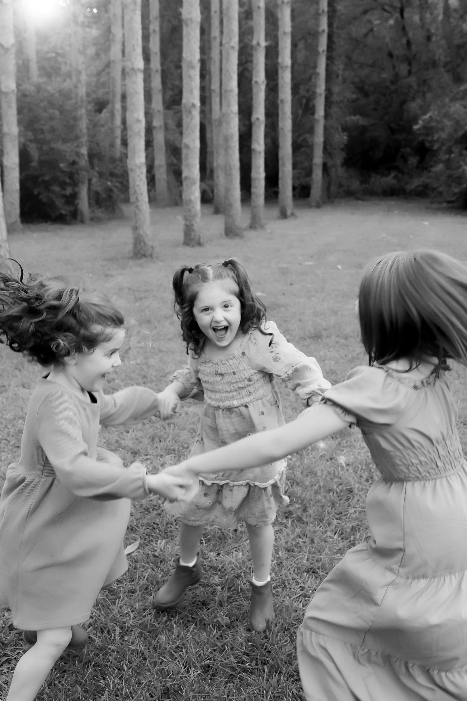 Three children in dresses playing a joyful game of ring-around-the-rosy in a forest clearing on a sunny day.