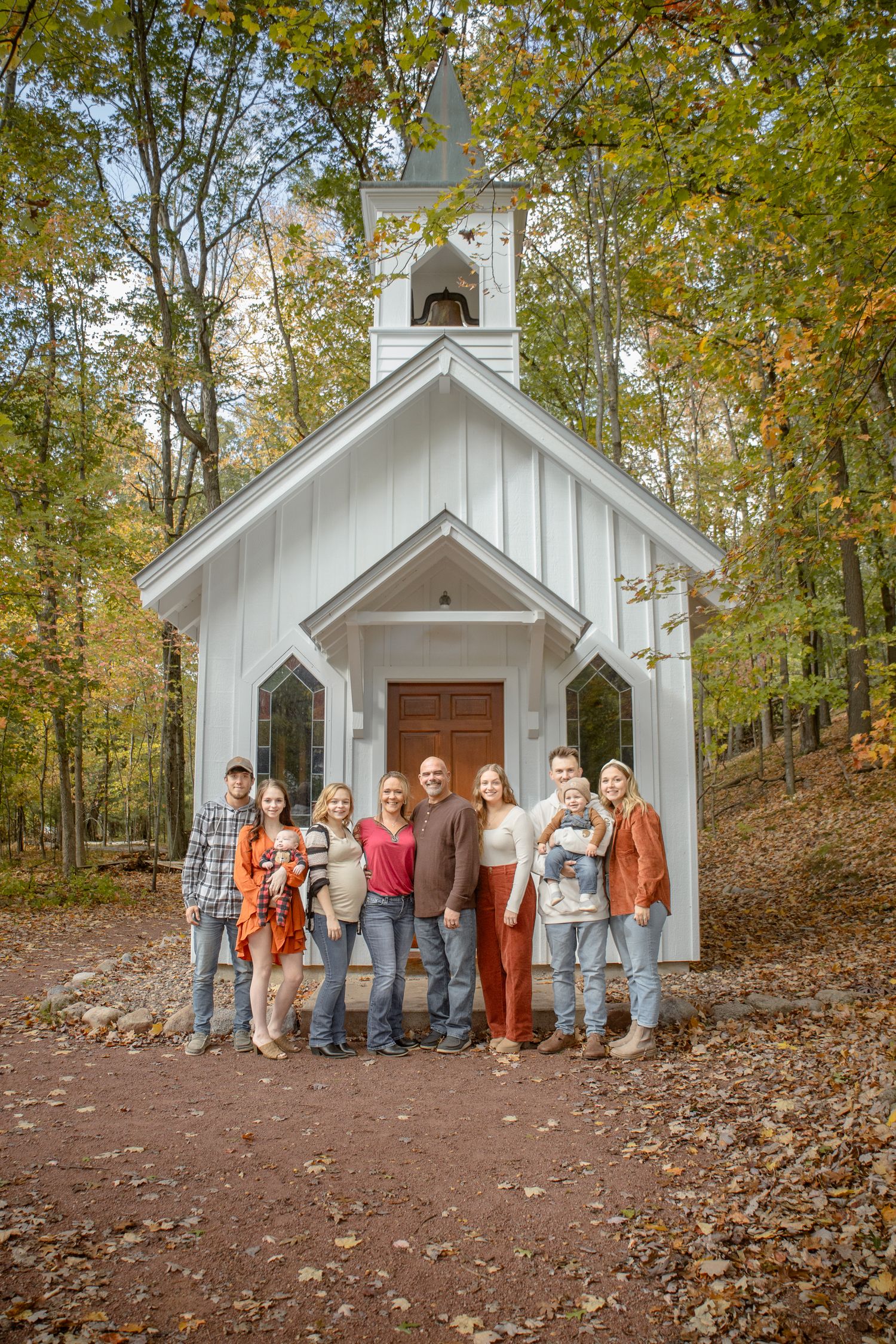 Family gathering posed in front of charming white chapel in autumn woods.