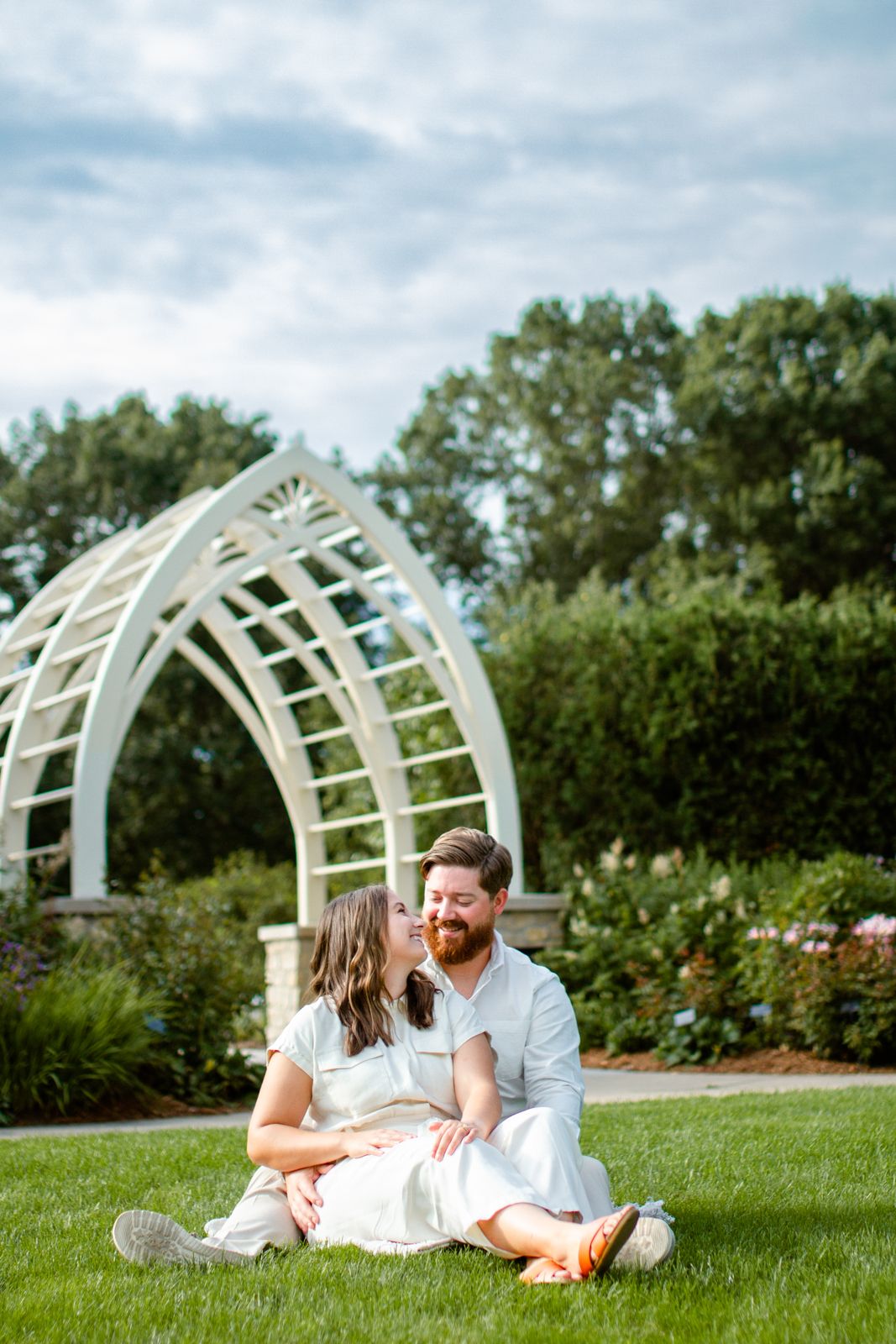 A couple relaxes on lawn near decorative white arched trellis in garden setting.