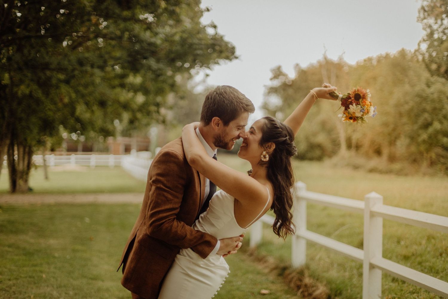 A couple shares a romantic kiss in a golden-lit field with white fencing during sunset.
