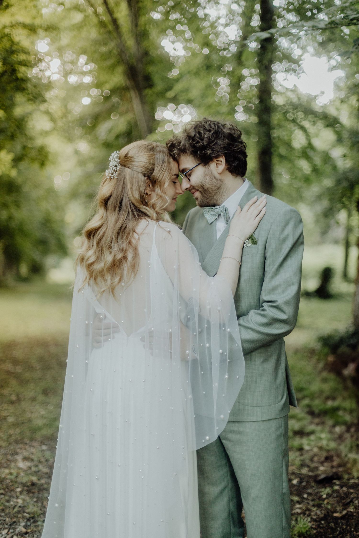 A romantic couple in sage green and white wedding attire share an intimate moment in a sunlit forest setting.