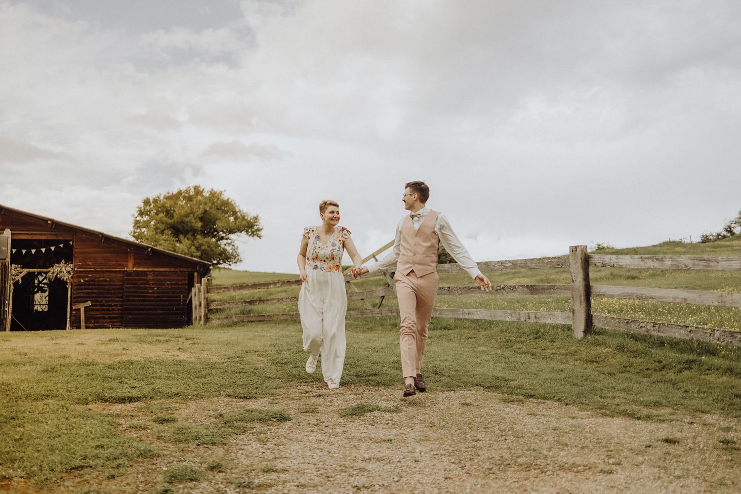 A couple in light-colored attire walks together on a grassy hillside during golden hour.