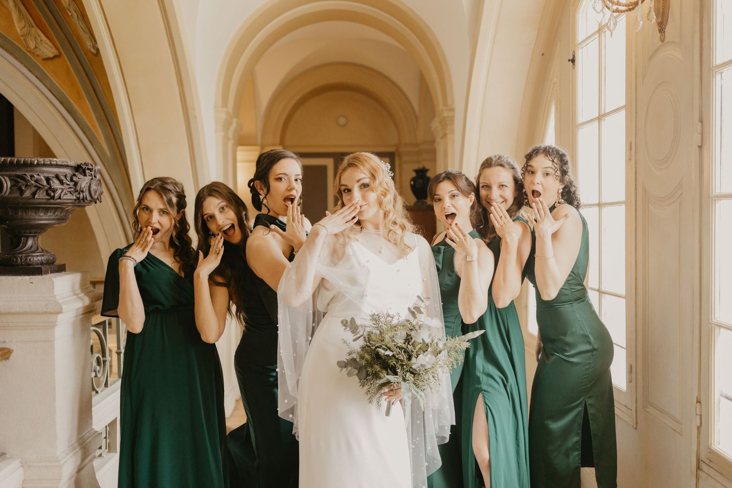 Bridesmaids in elegant emerald green dresses gather with the bride in a bright, sunlit hallway for wedding photos.
