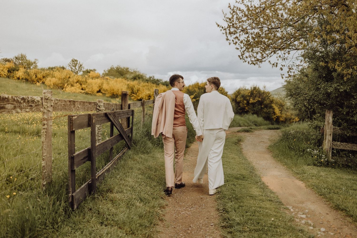 Two people walk hand in hand down a winding dirt path surrounded by lush green countryside on a sunny autumn day.