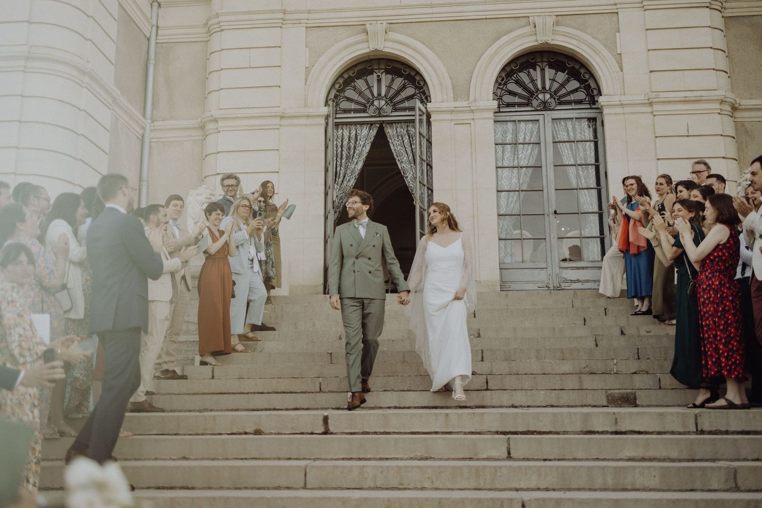 A couple descends stone steps outside a historic building with arched doorways while guests line both sides of the staircase.