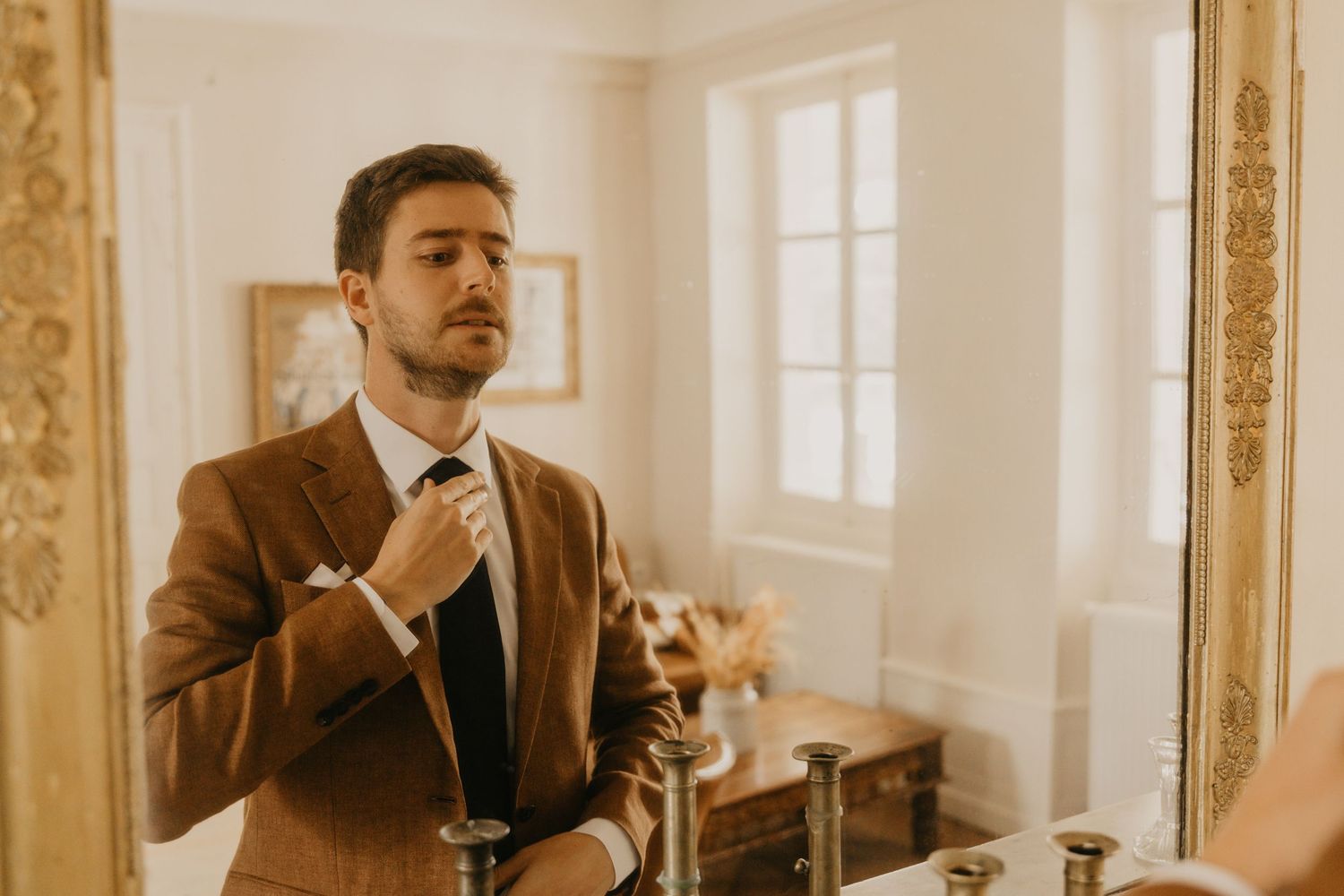 A groom adjusts his tie while getting ready in an elegant room with ornate mirrors and warm lighting.