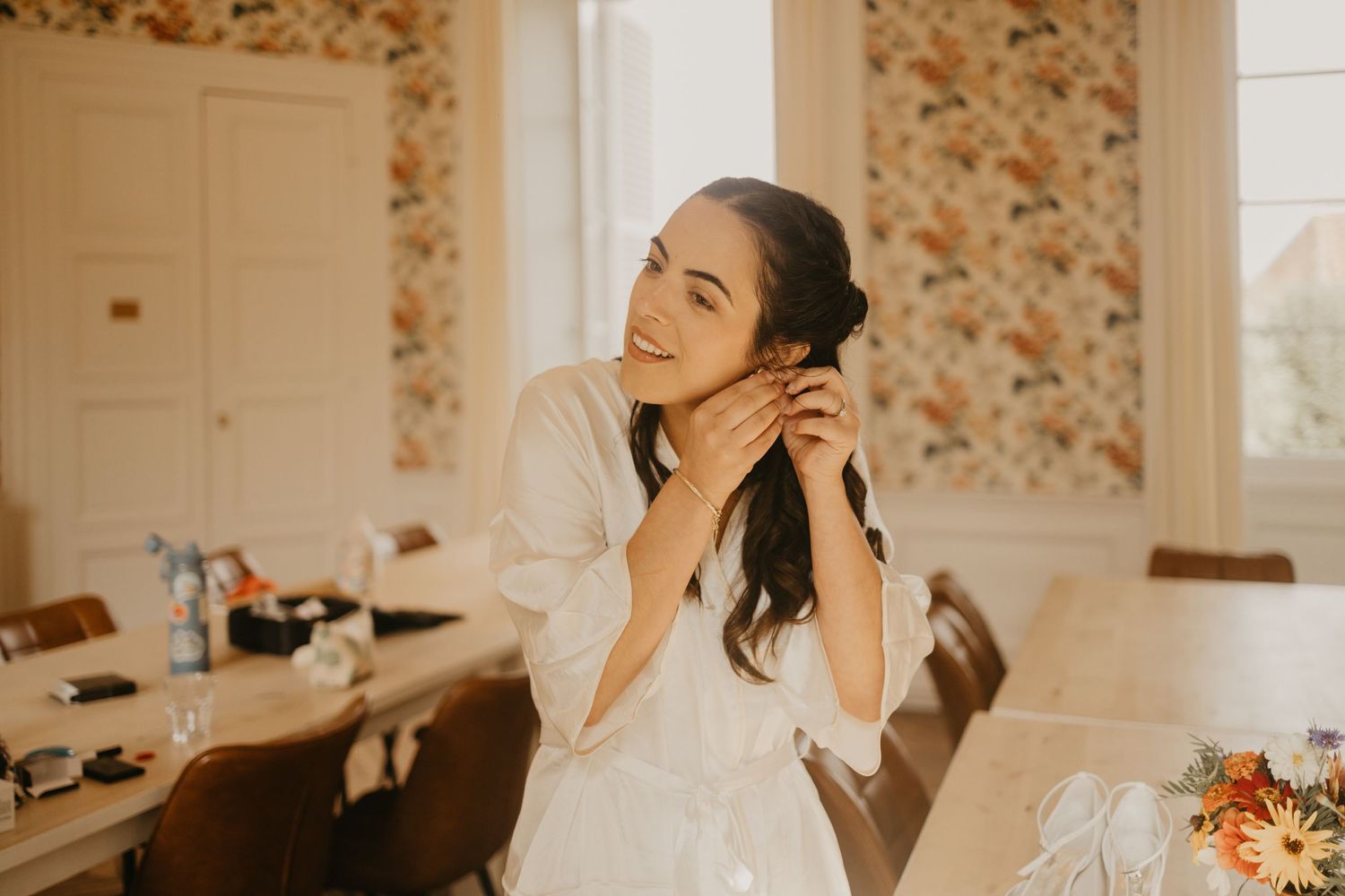 A person laughs joyfully while sitting in a bright room with floral wallpaper and natural lighting.
