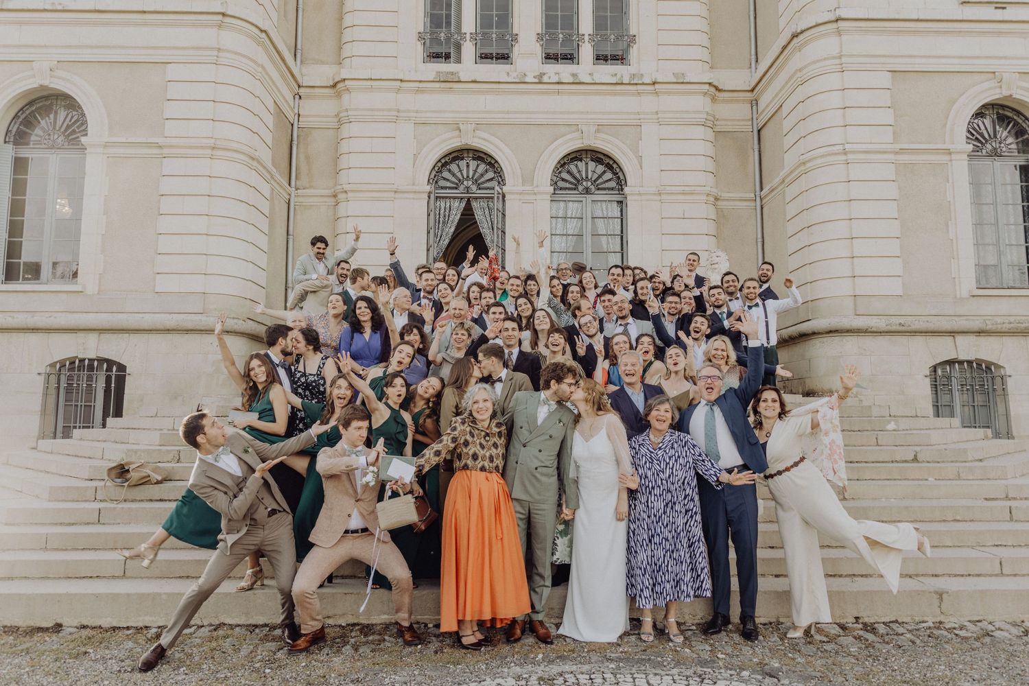 Large wedding party poses for a group photo outside an elegant stone building with classical architecture.