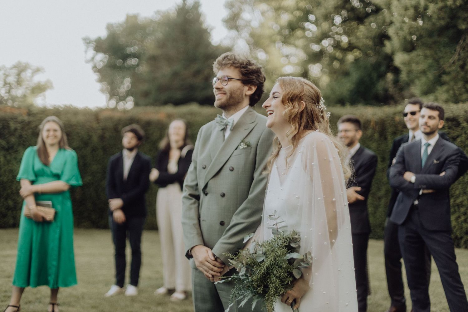 Wedding party stands outdoors with bridesmaids in emerald green dresses and groomsmen in dark suits.