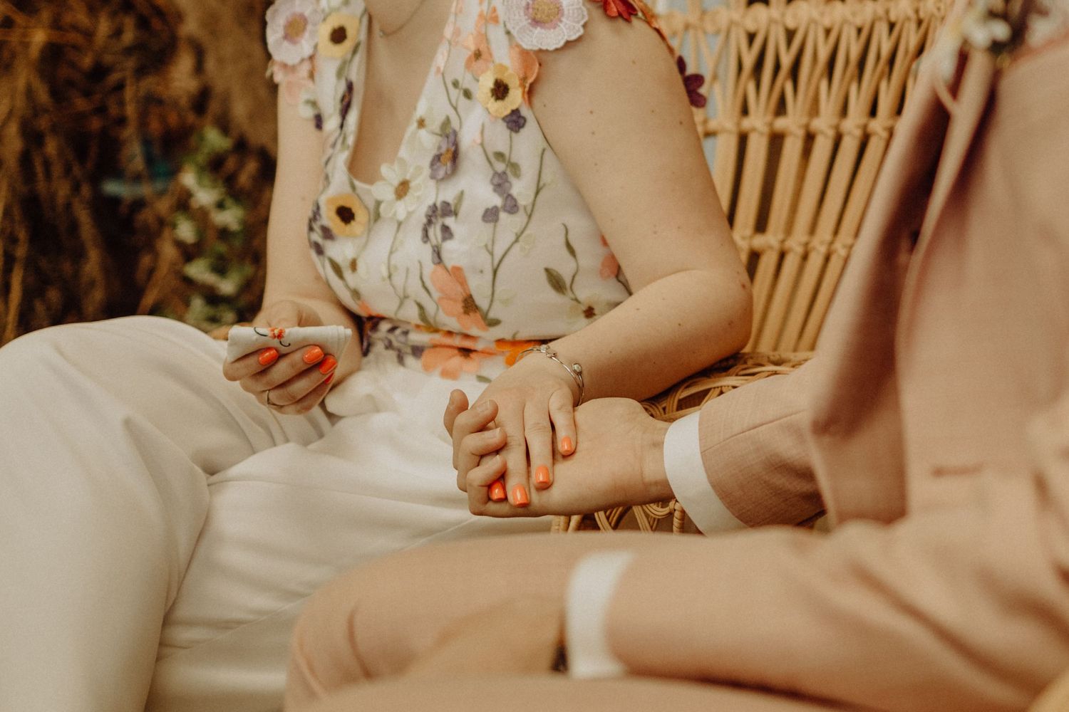 Two people sitting in a wicker chair hold hands intimately in warm sunlight wearing floral and neutral colored clothing.