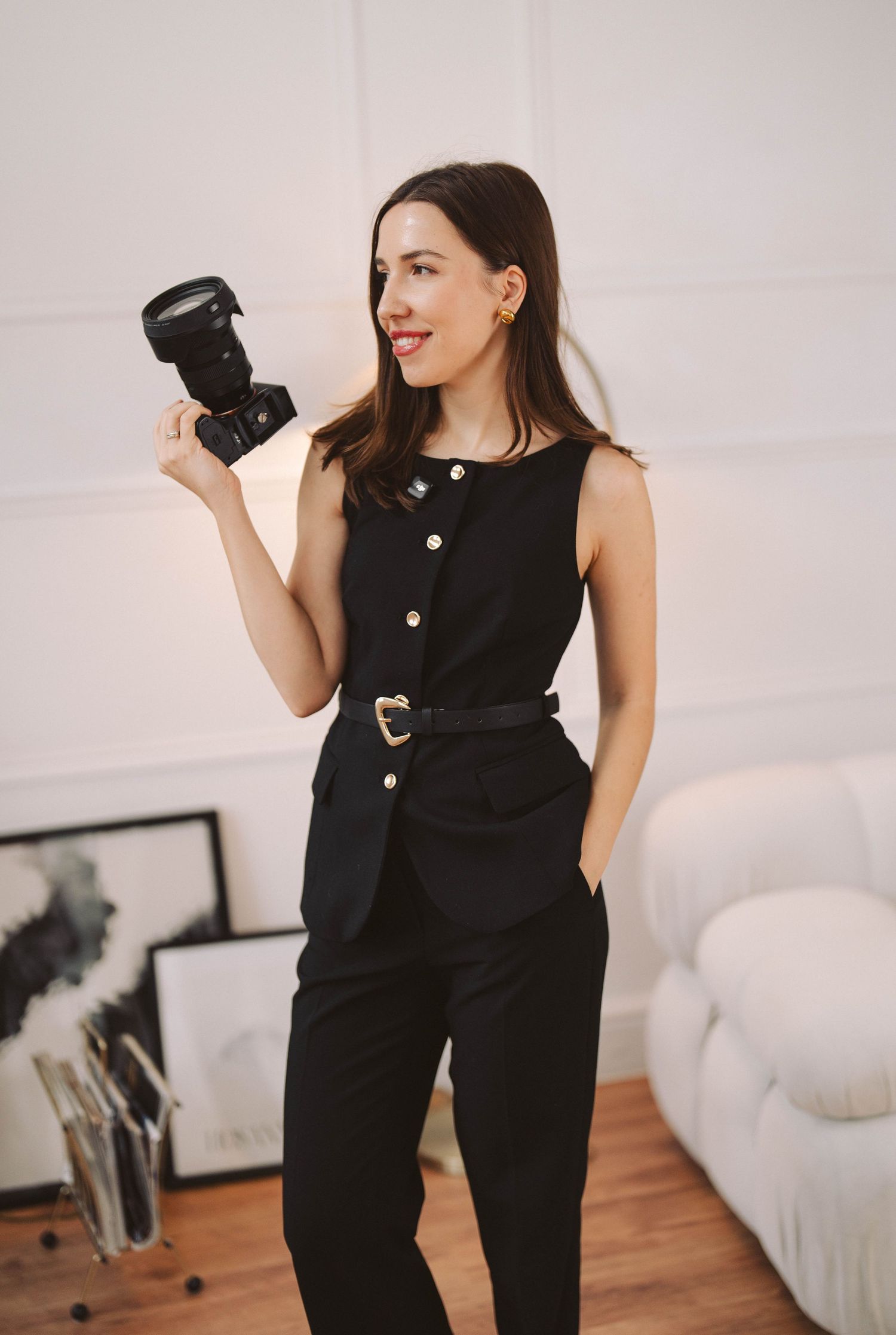 Professional photographer in elegant black jumpsuit holds DSLR camera while posing in minimalist studio setting.