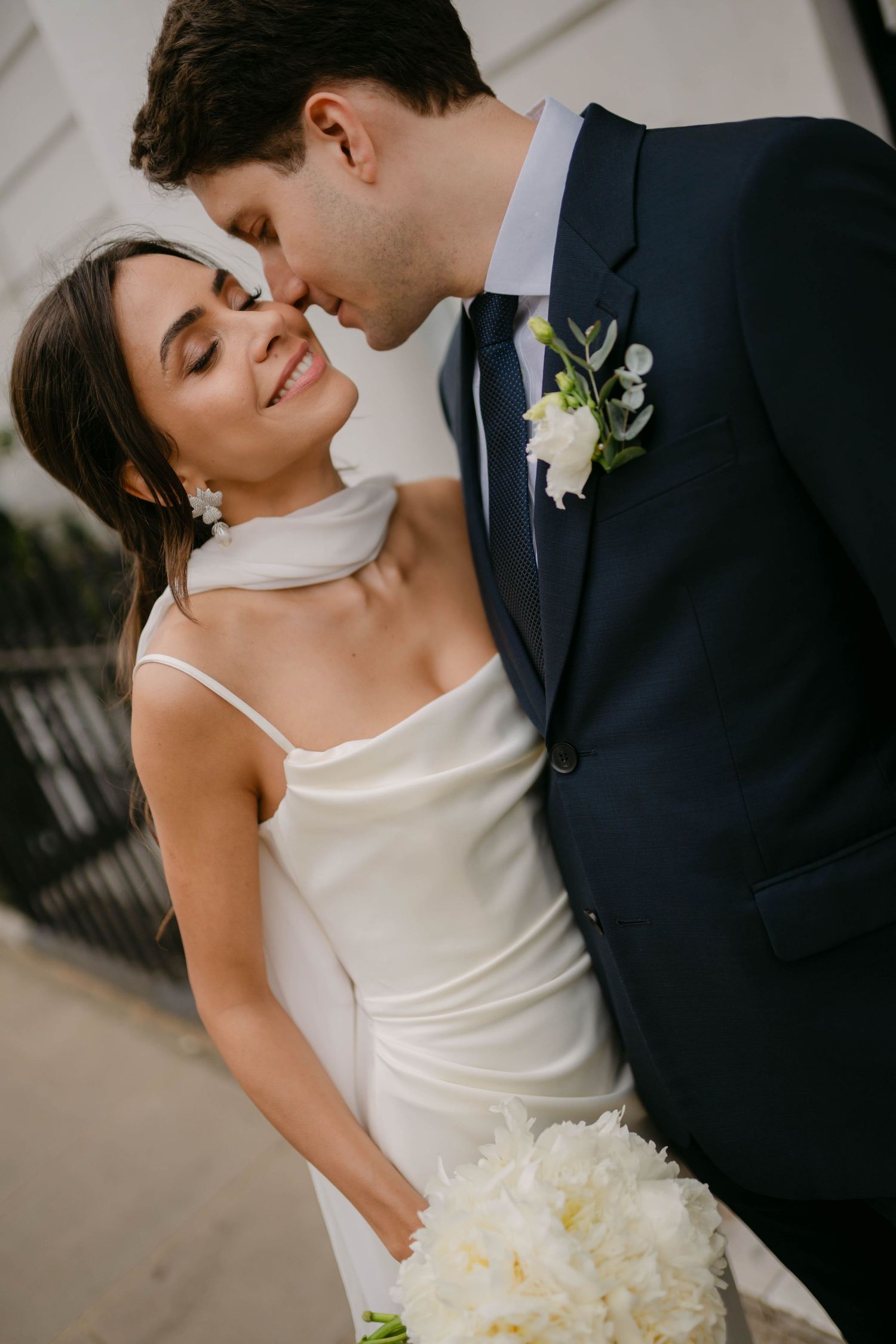 Elegant bride in white gown holds bouquet while sharing intimate moment with groom in navy suit.