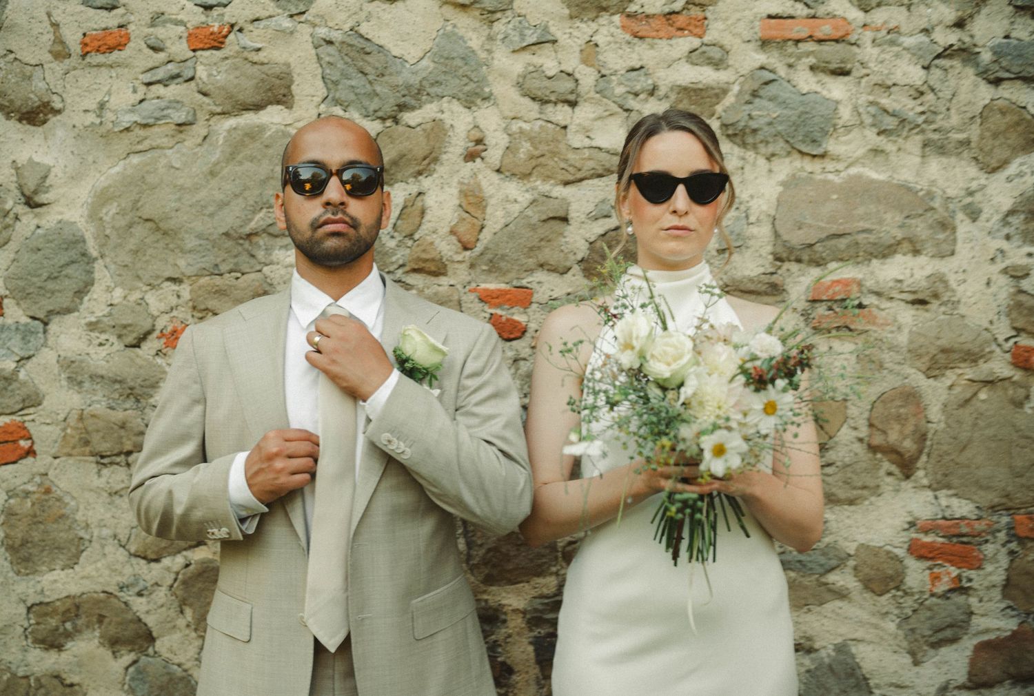 Wedding party posing casually against rustic stone wall wearing sunglasses.