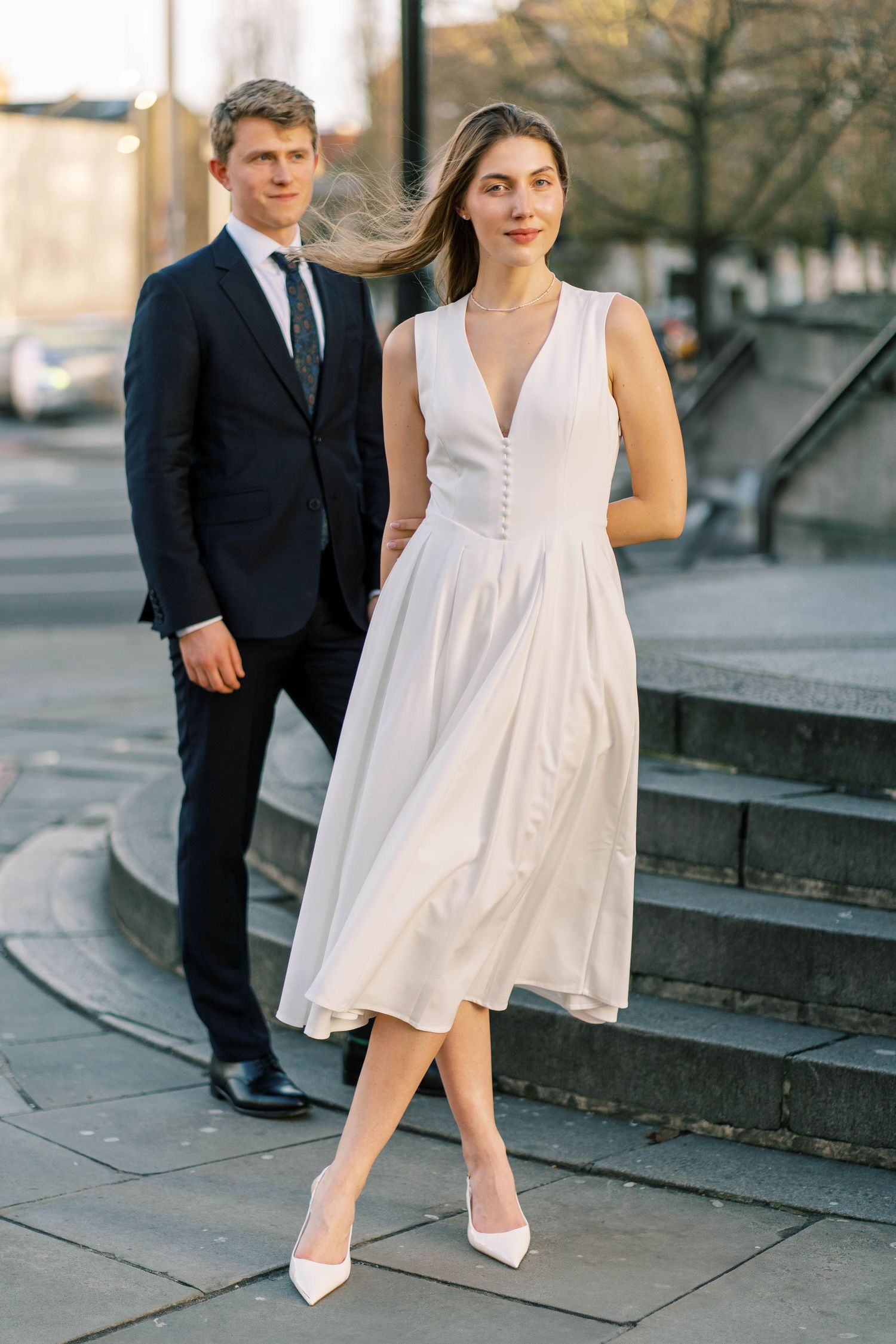A couple poses on stone steps, with one wearing a white dress and the other in a dark suit.