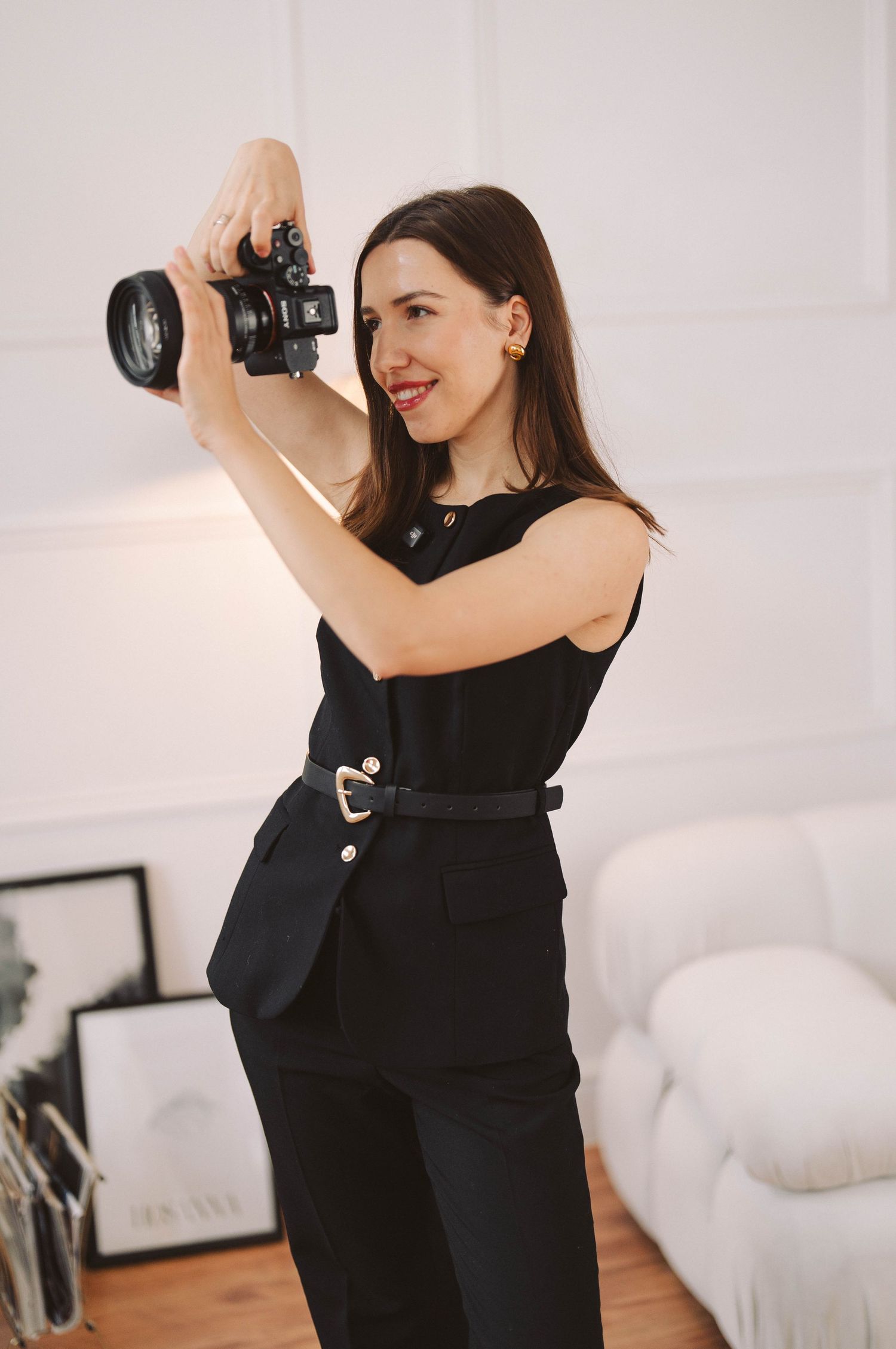 Professional photographer in elegant black jumpsuit holds camera while shooting in bright studio interior.